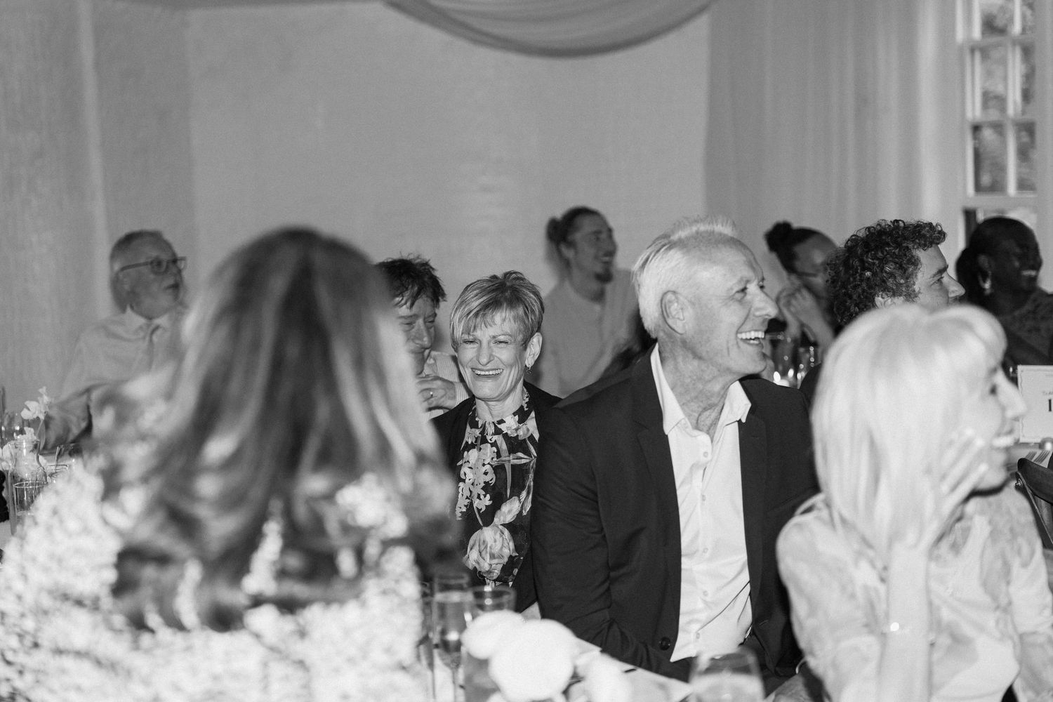 Black and white wedding reception photo of smiling guests seated at tables during a celebratory dinner event.