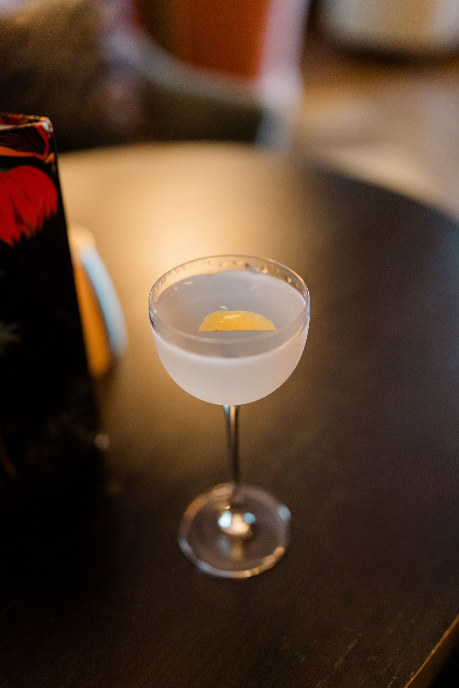 A frosted coupe cocktail glass with a lemon twist garnish on a dark wooden bar table.