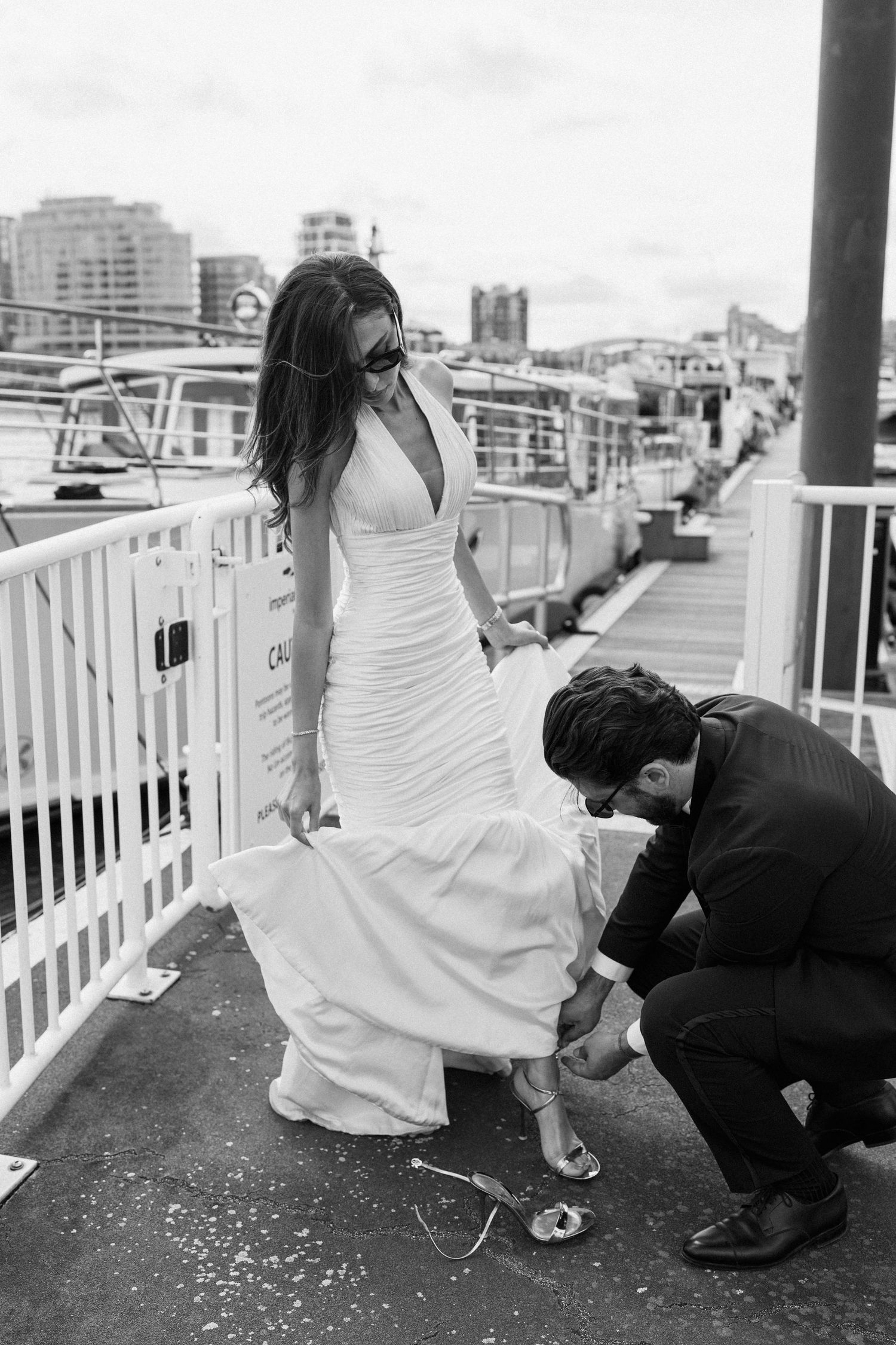 Black and white photo of groom fixing bride's shoe on a marina dock, bride in elegant white gown wearing sunglasses.