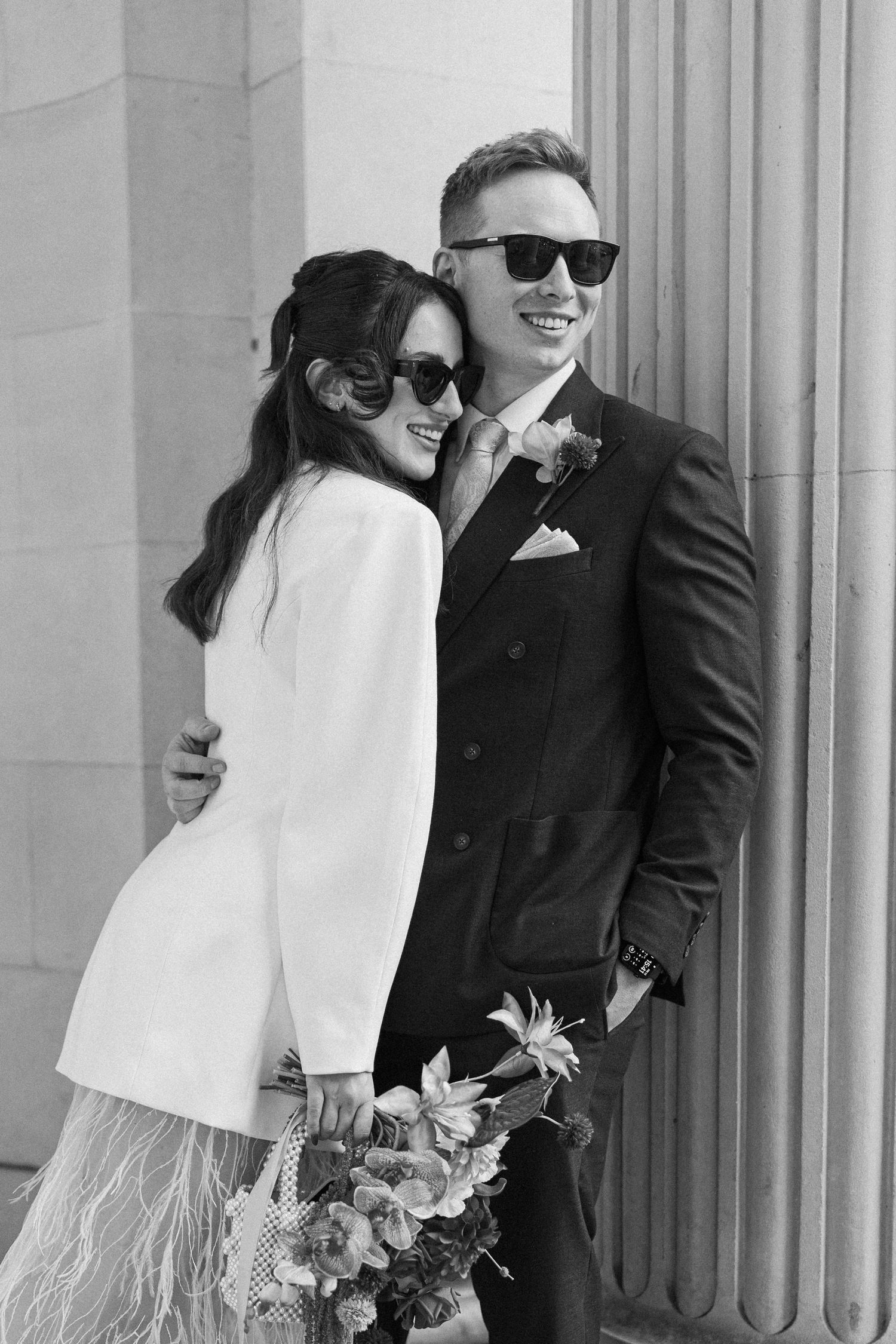 Black and white photo of stylish newlyweds wearing sunglasses posing against stone columns.