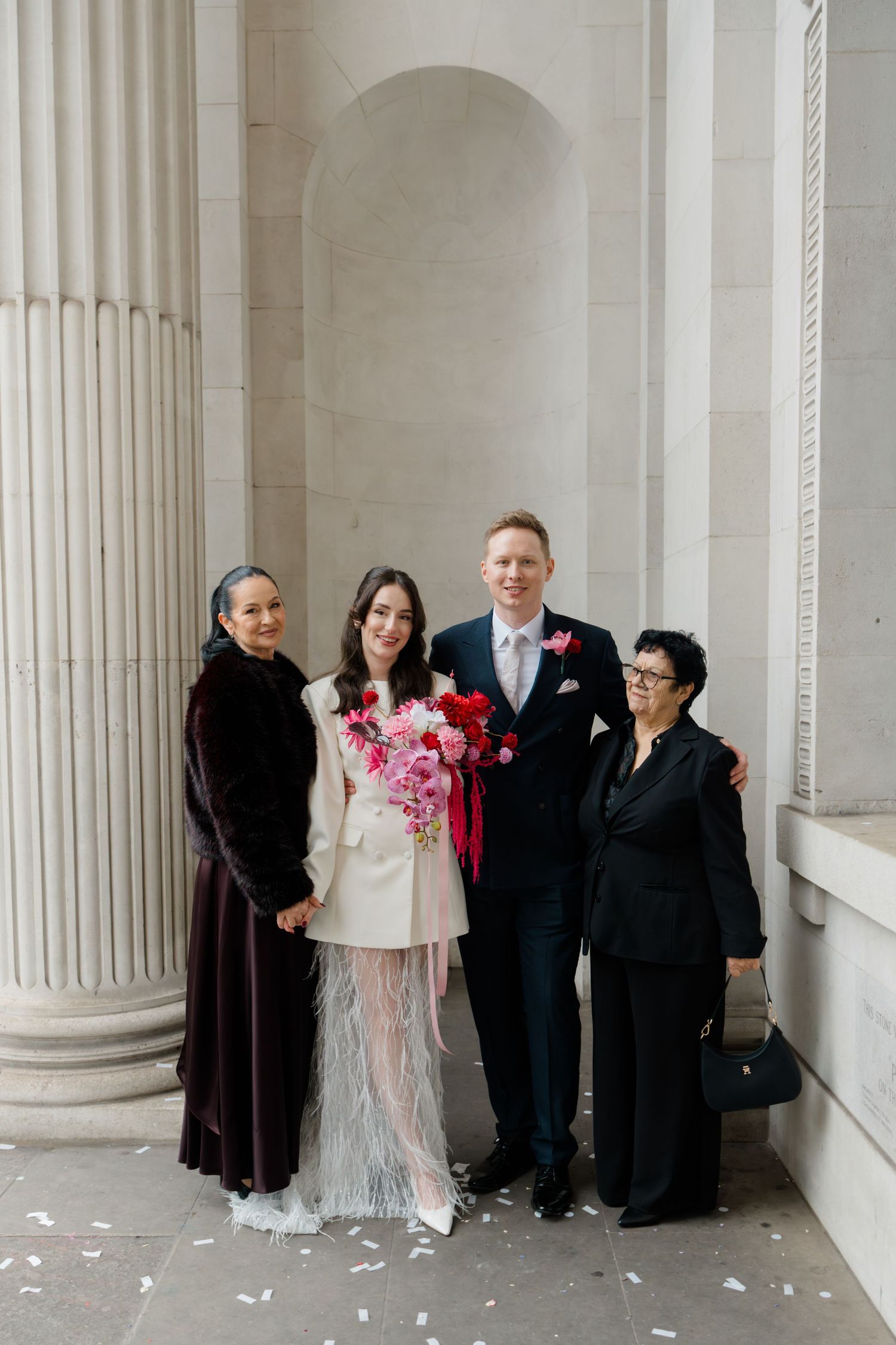 Bride and groom pose with two female family members outside stone columned wedding venue.