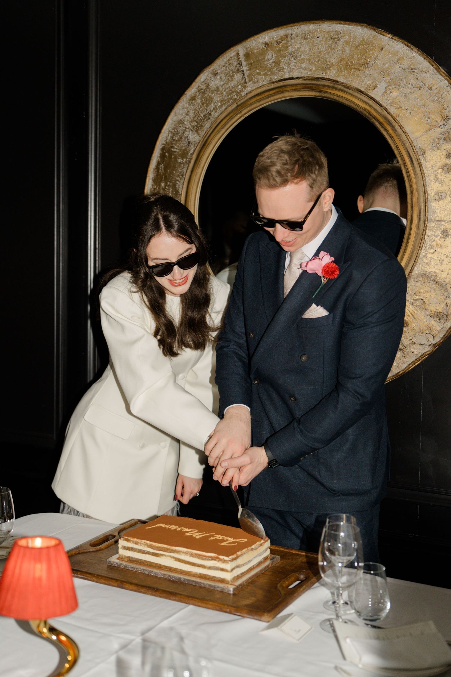 Newlyweds wearing sunglasses cut tiered wedding cake together at elegant dark-walled reception.