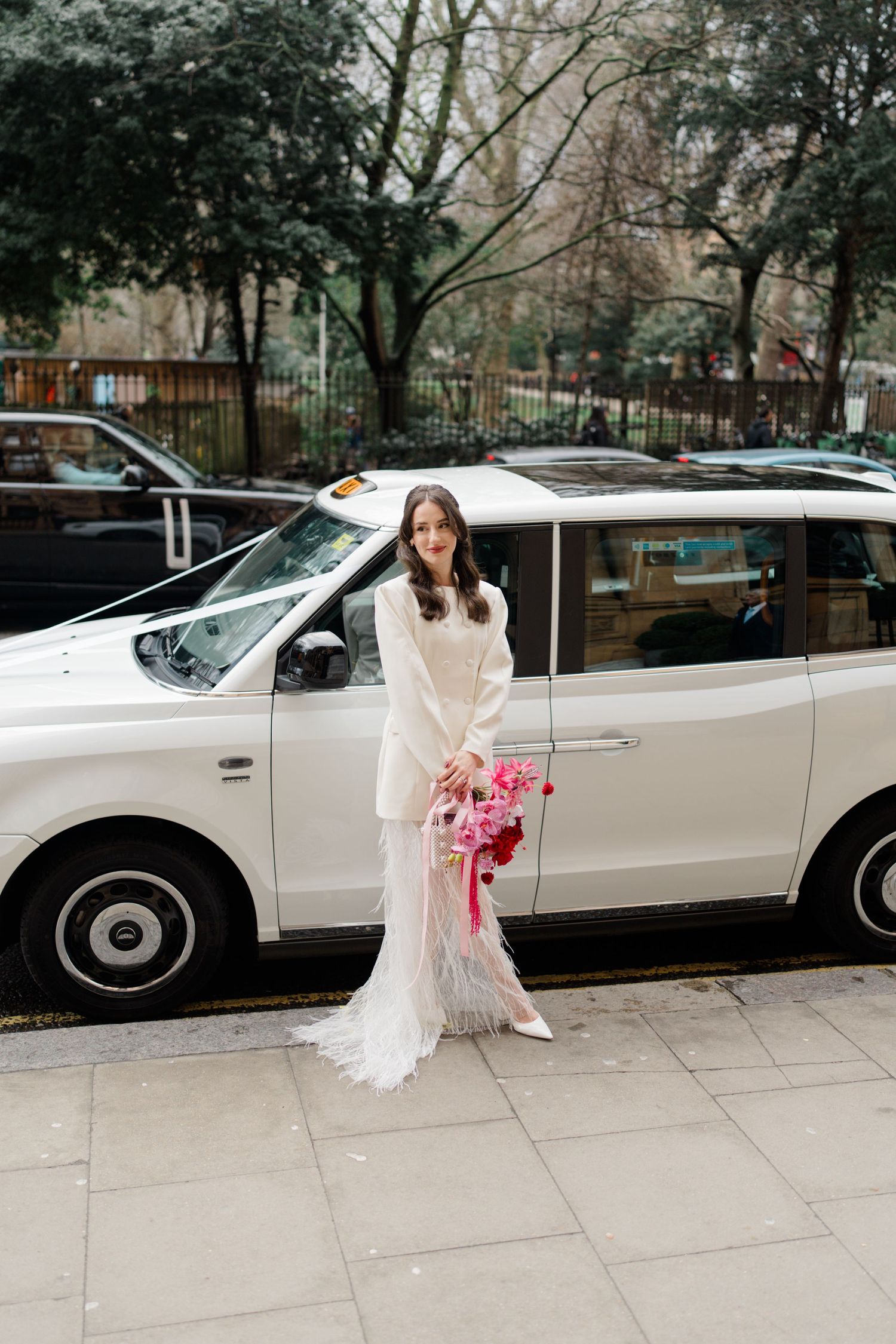 Bride in white sweater and feather skirt holding pink bouquet beside white London taxi cab.