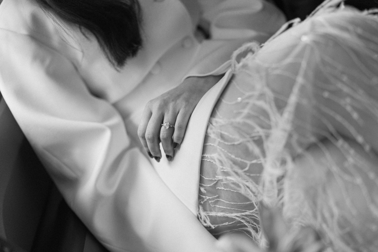 Close-up black and white photo of bride in white blazer with feather detail and wedding ring resting hand on lap.