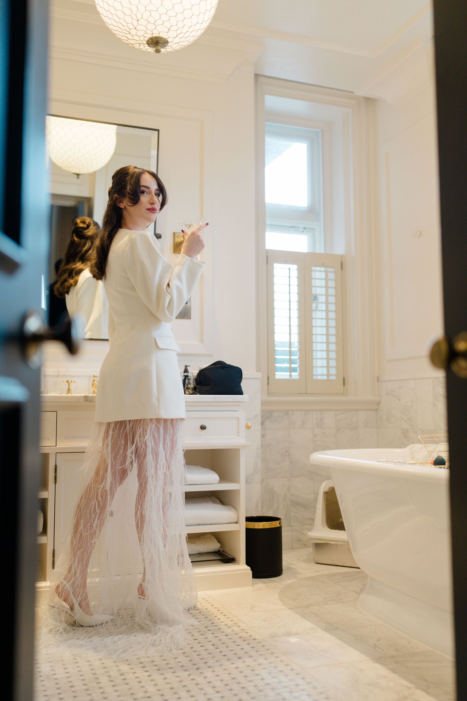 Elegant bride in white blazer and feathered skirt applies makeup in a bright marble hotel bathroom.