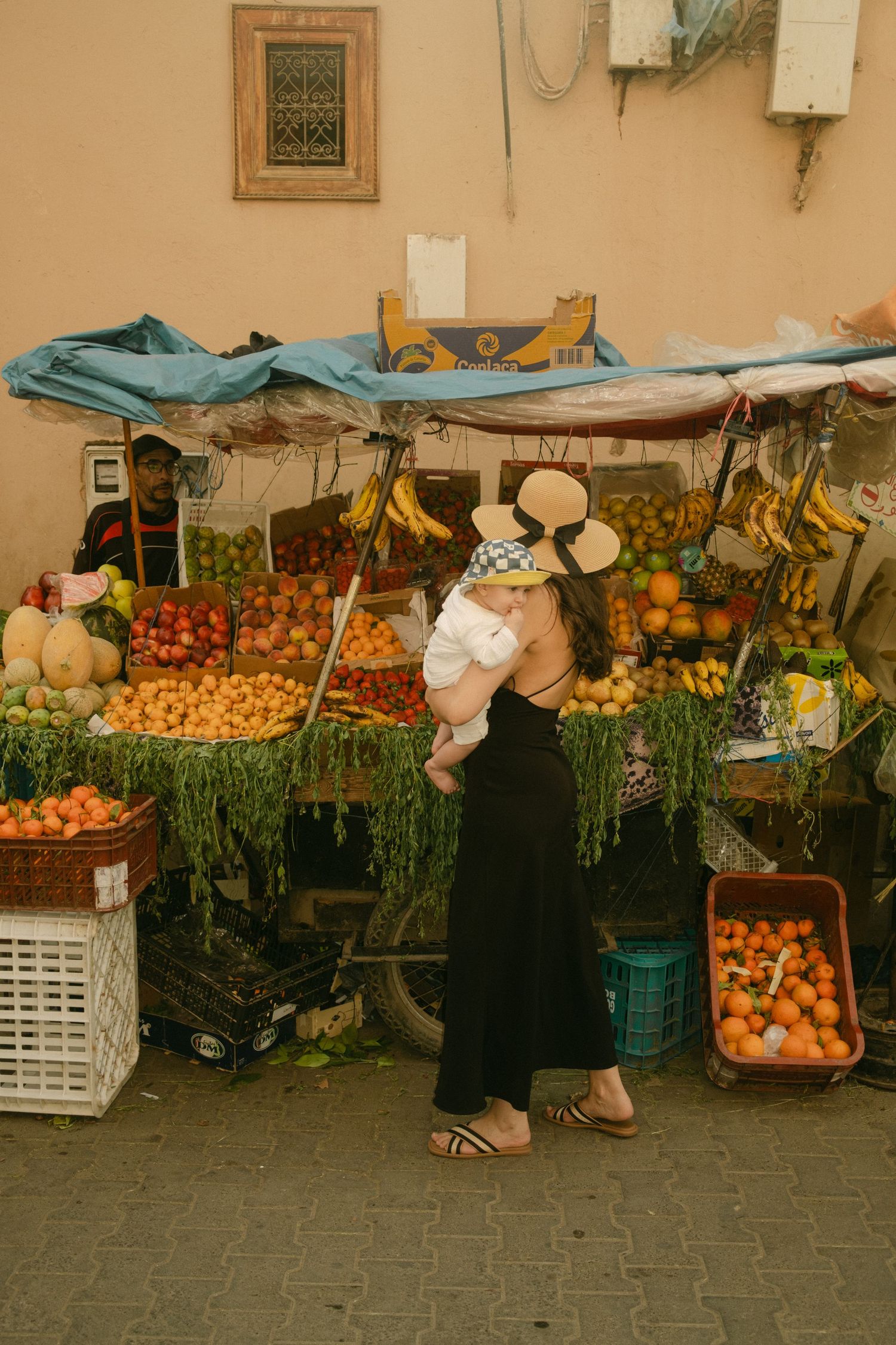 Woman holding baby at colorful outdoor fruit market stall with fresh produce display.