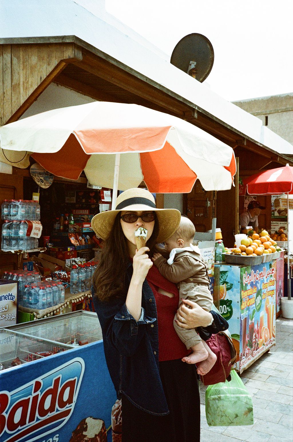 Woman in sun hat and sunglasses holds baby under colorful market umbrella near outdoor fruit and beverage stalls.