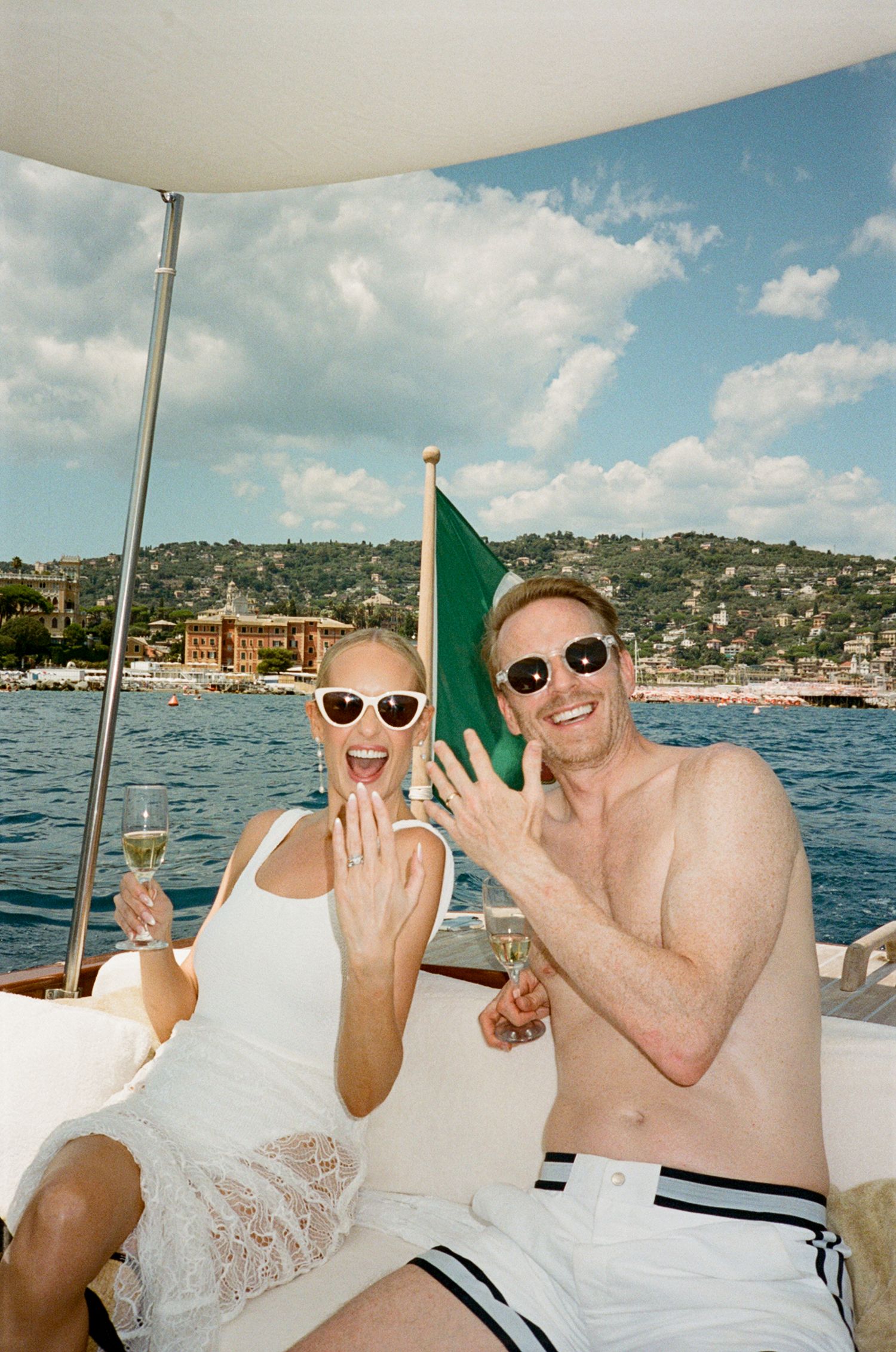 Happy couple on a luxury boat showing engagement ring, holding champagne glasses with scenic coastal town backdrop.