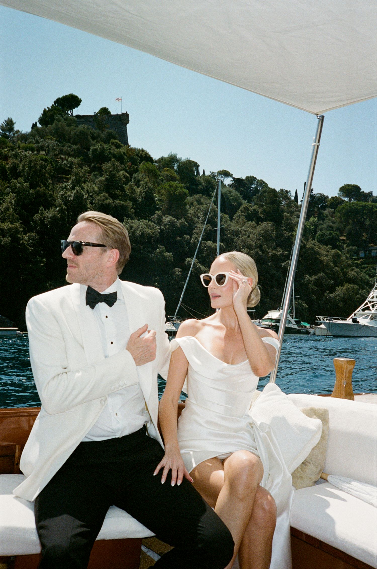 Elegant couple in wedding attire on a luxury boat in Portofino, Italy, wearing sunglasses on a sunny day.