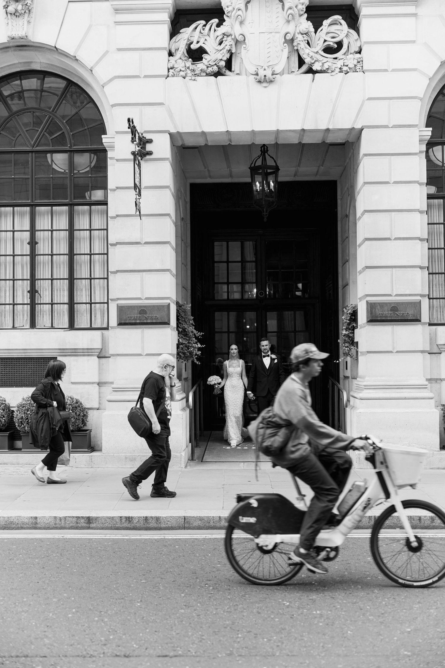 Black and white street scene with a cyclist passing by a grand building entrance during a wedding.