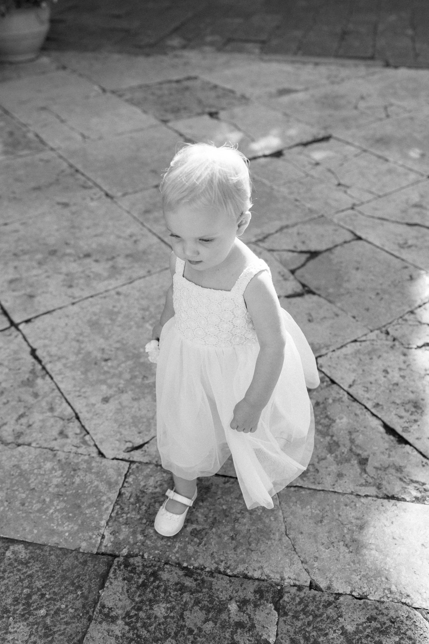 Small child in white dress stands on stone patio in black and white photograph.