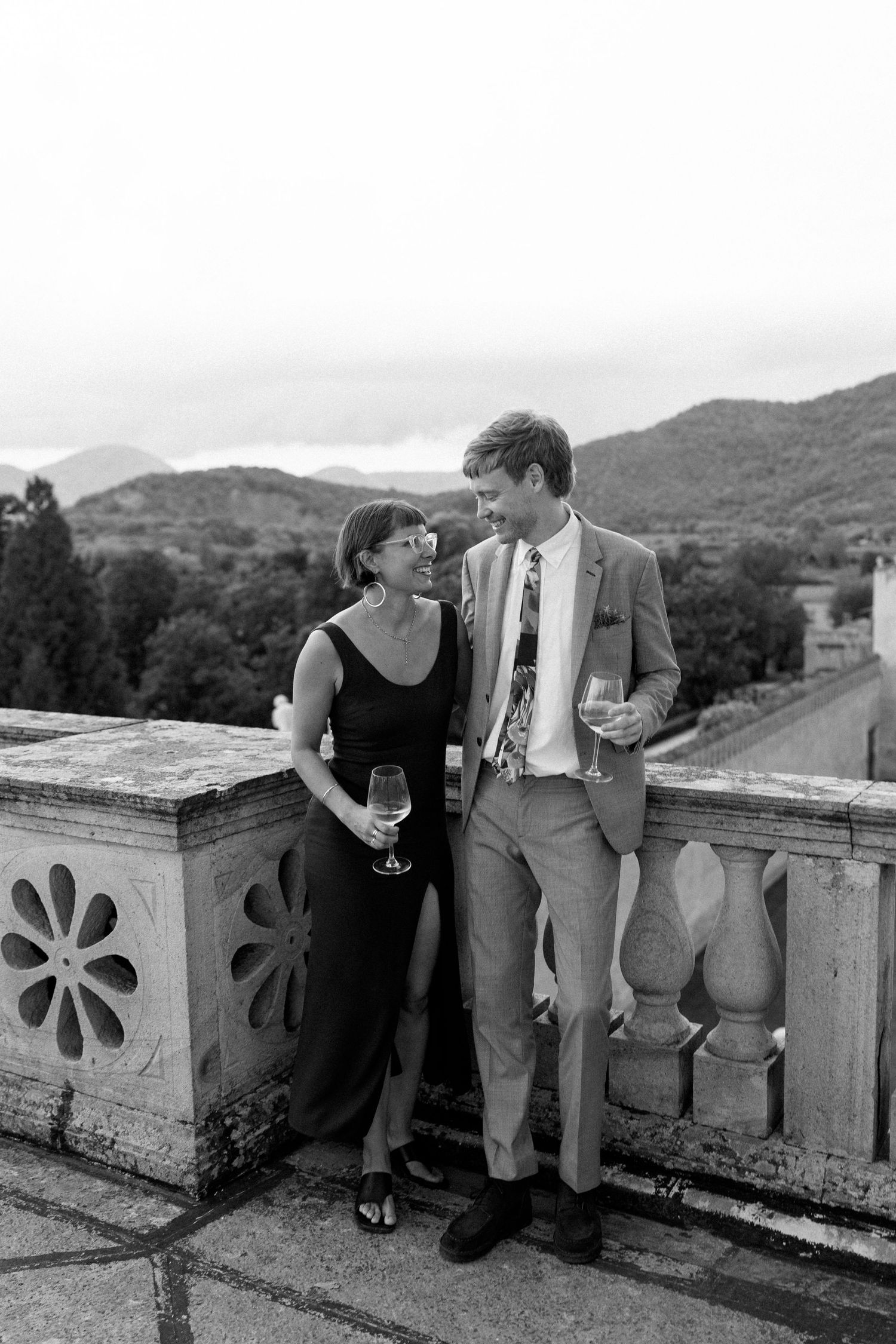 A couple enjoying drinks while leaning against a decorative stone balustrade with mountain views.