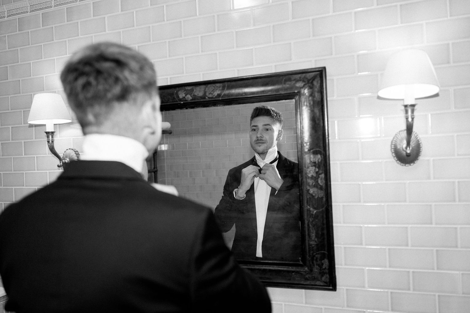 Black and white photo of someone adjusting their tie in a mirror with brick wall background.