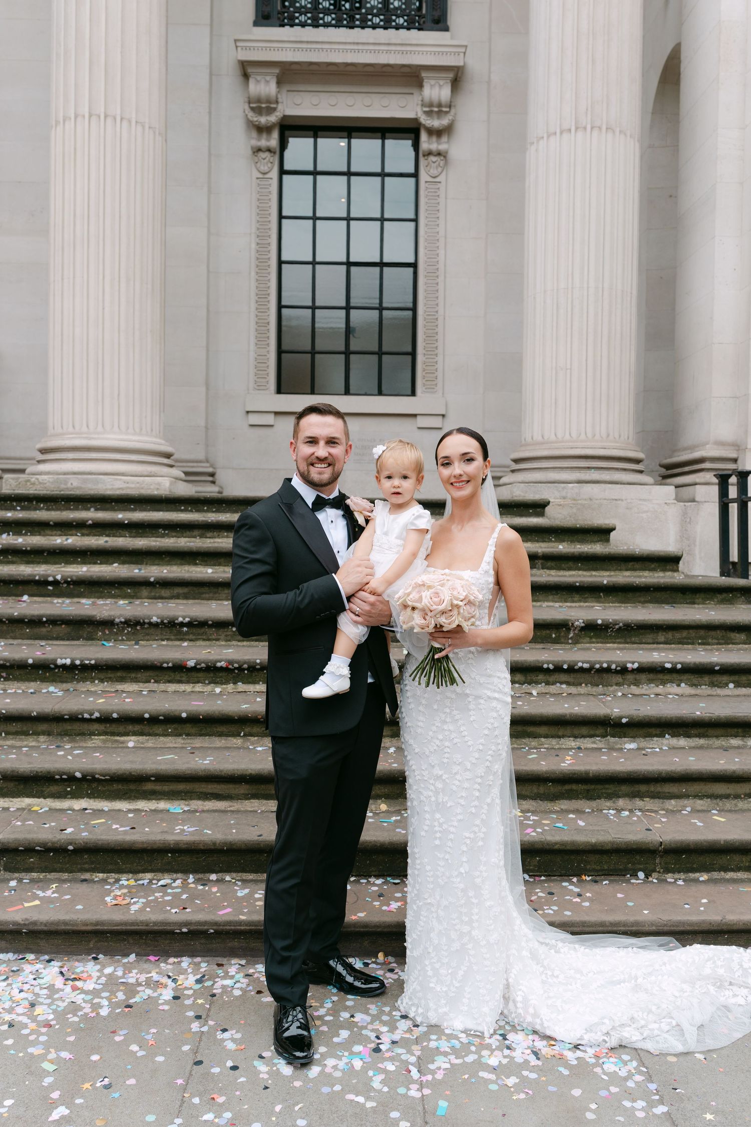 Wedding party poses on stone steps of a classical building with scattered flower petals.