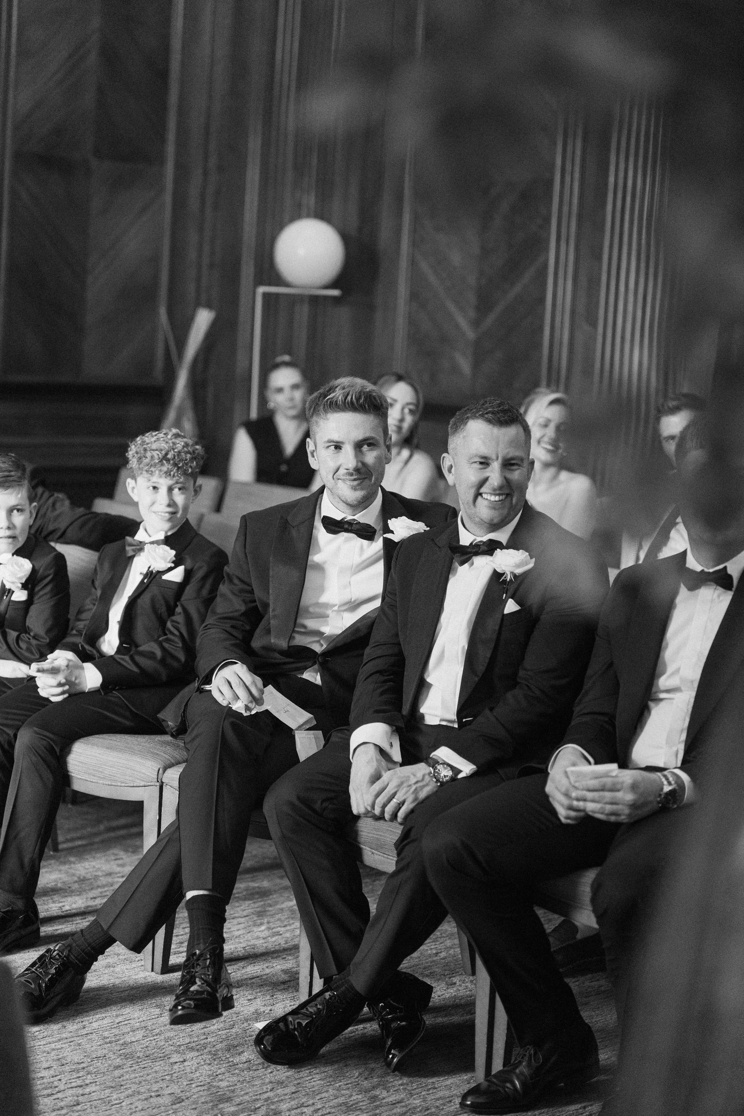 Groomsmen in black tuxedos sitting casually against a wall during wedding festivities.