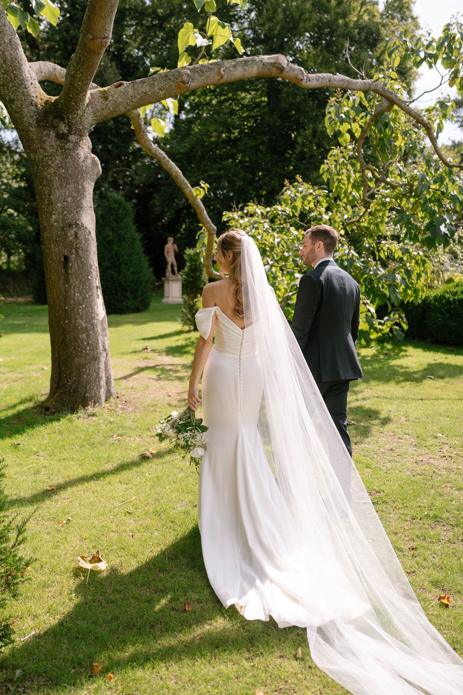 Wedding couple poses under large trees in a sunlit garden setting.