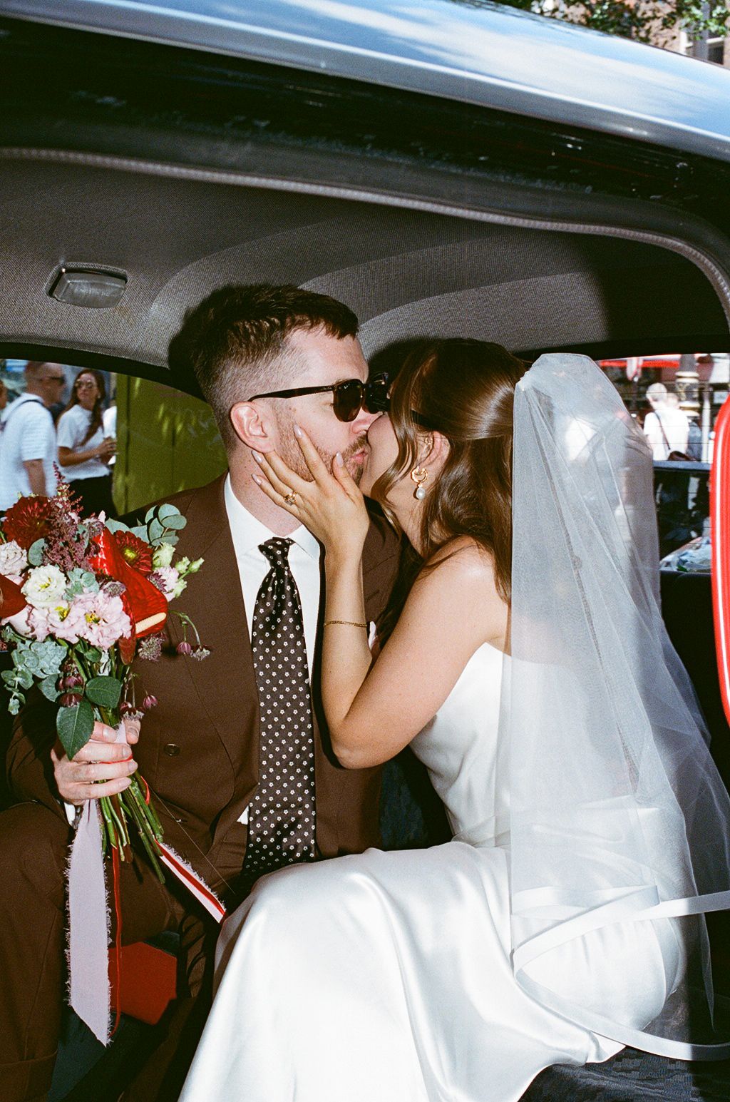 Newlyweds sharing a kiss in a car, groom in brown suit holding floral bouquet, bride in white dress with veil.
