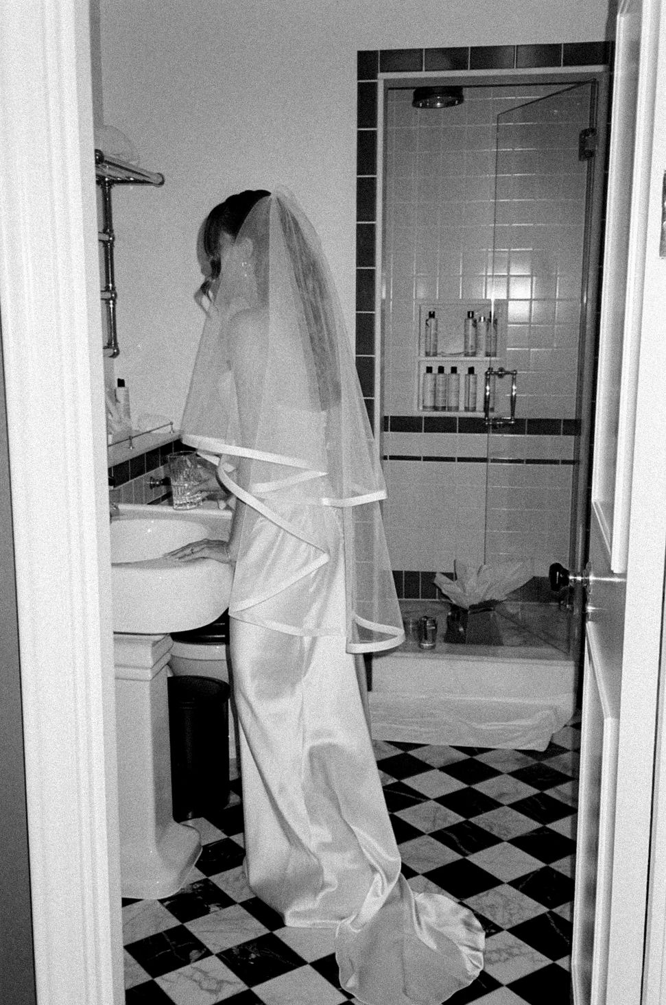 Bride in white wedding dress and veil standing at bathroom sink, black and white photo with checkered floor.