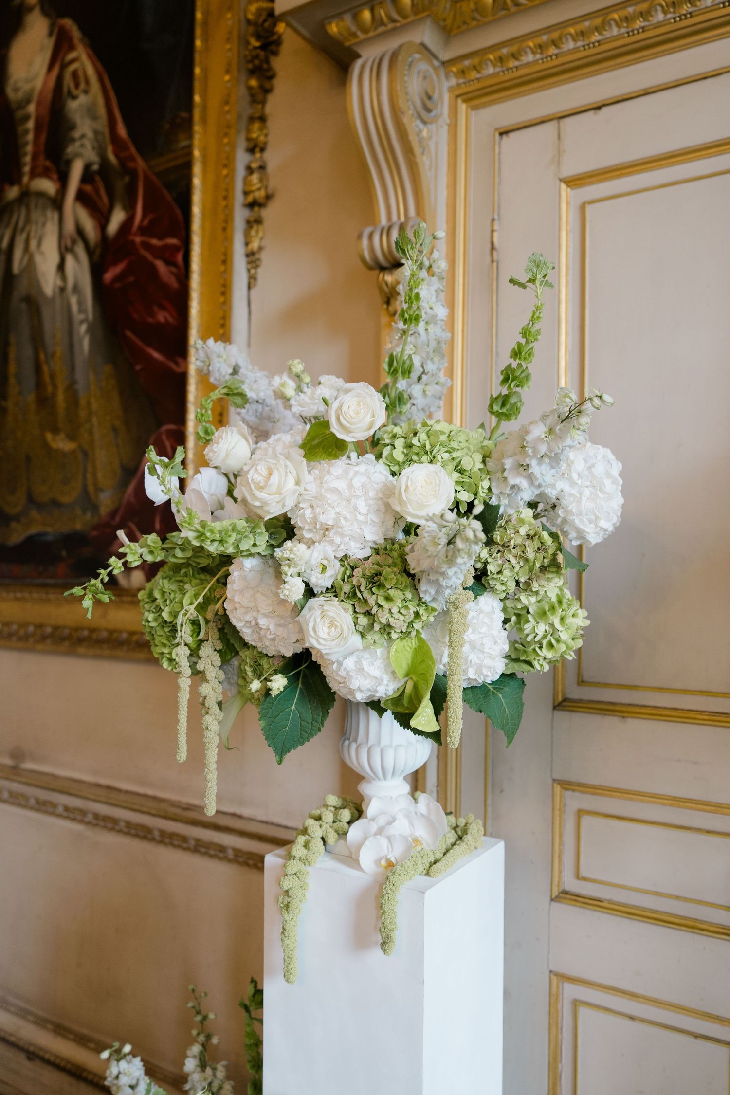 Lush white floral arrangement with roses and hydrangeas in a white urn on a pedestal in an elegant ballroom.