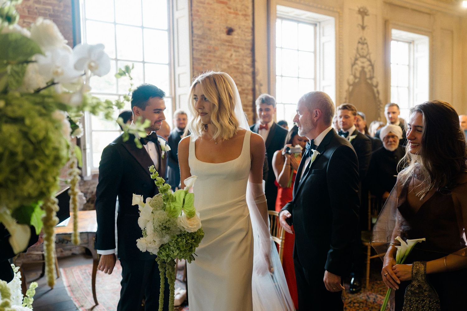 Bride in white slip dress walks down aisle with bouquet at elegant brick wedding venue.