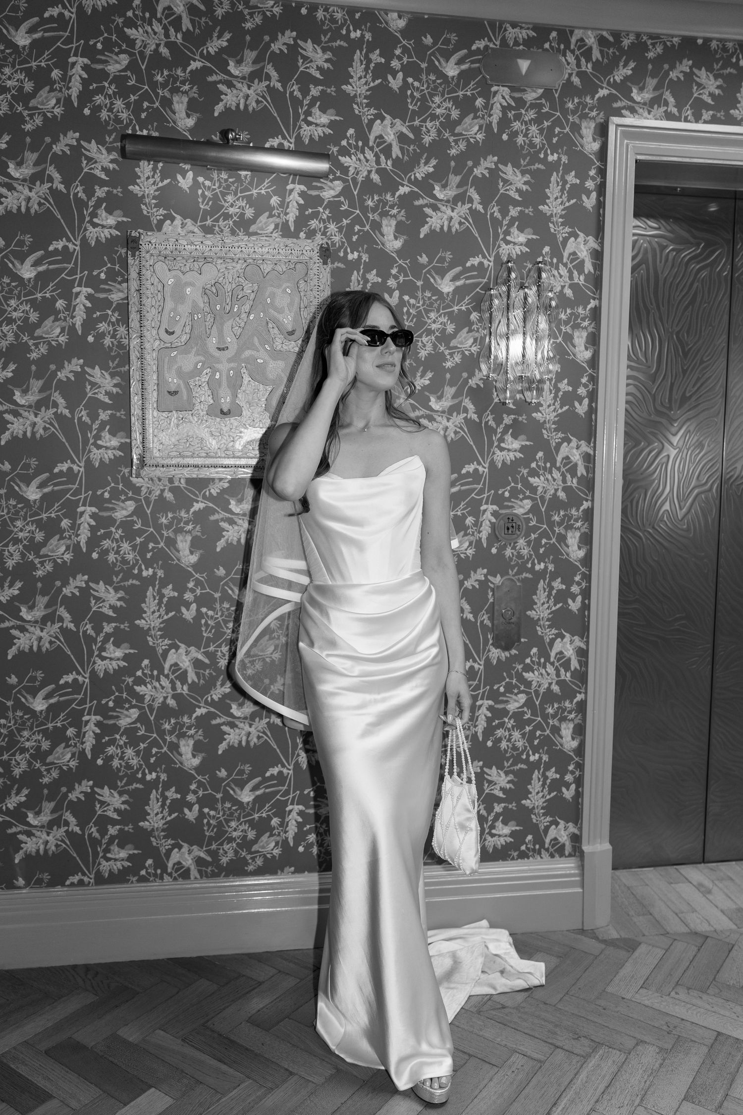 Black and white photo of bride in satin gown and sunglasses posing by ornate floral wallpaper.