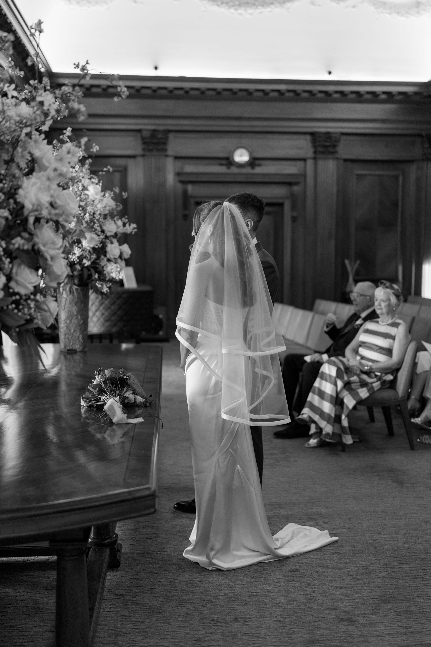 Black and white photo of bride and groom during wedding ceremony in elegant wood-paneled room.
