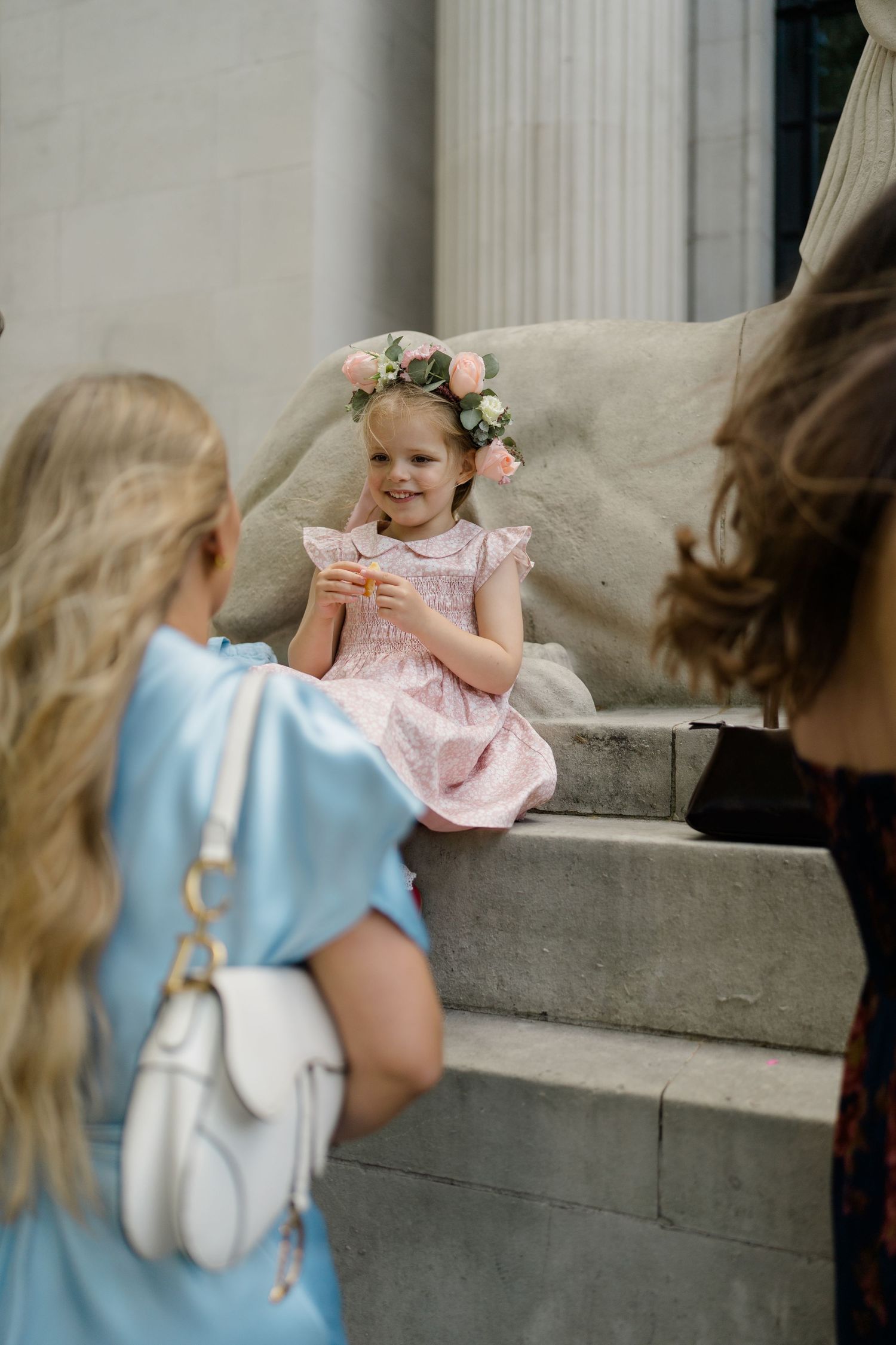 Smiling flower girl in pink dress and floral crown sits on stone steps at wedding.