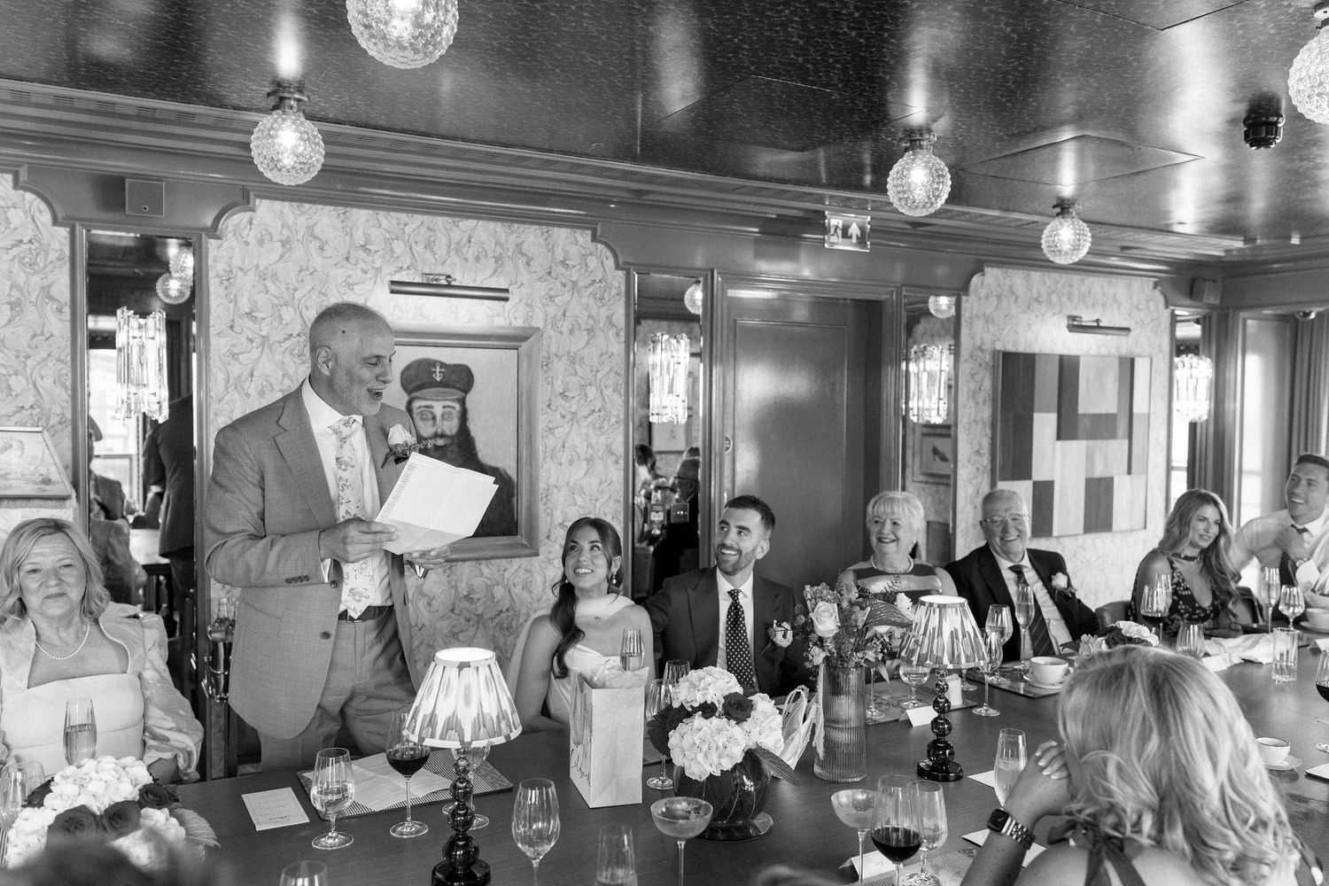 Wedding guest delivers speech at long dining table in ornate black and white restaurant setting.