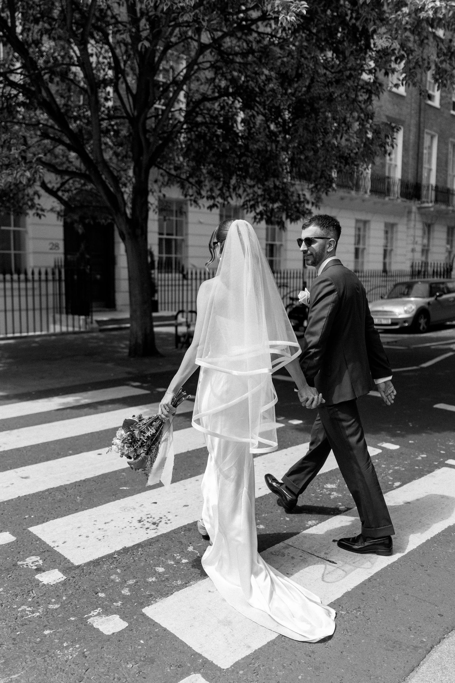 Black and white photo of bride and groom crossing a zebra crossing on a sunny city street.