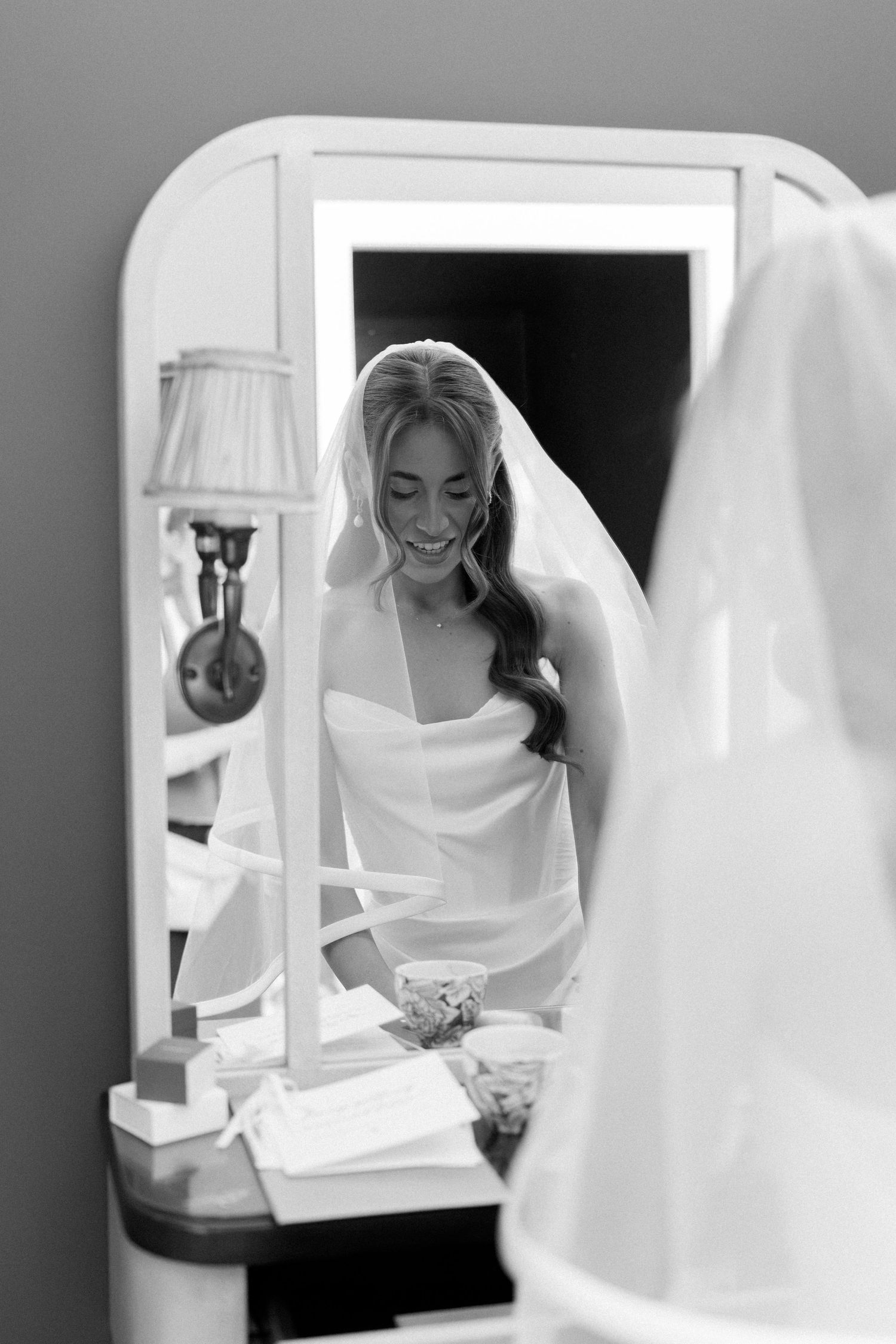 Black and white photo of a smiling bride in a strapless gown and veil, reflected in a lighted vanity mirror.