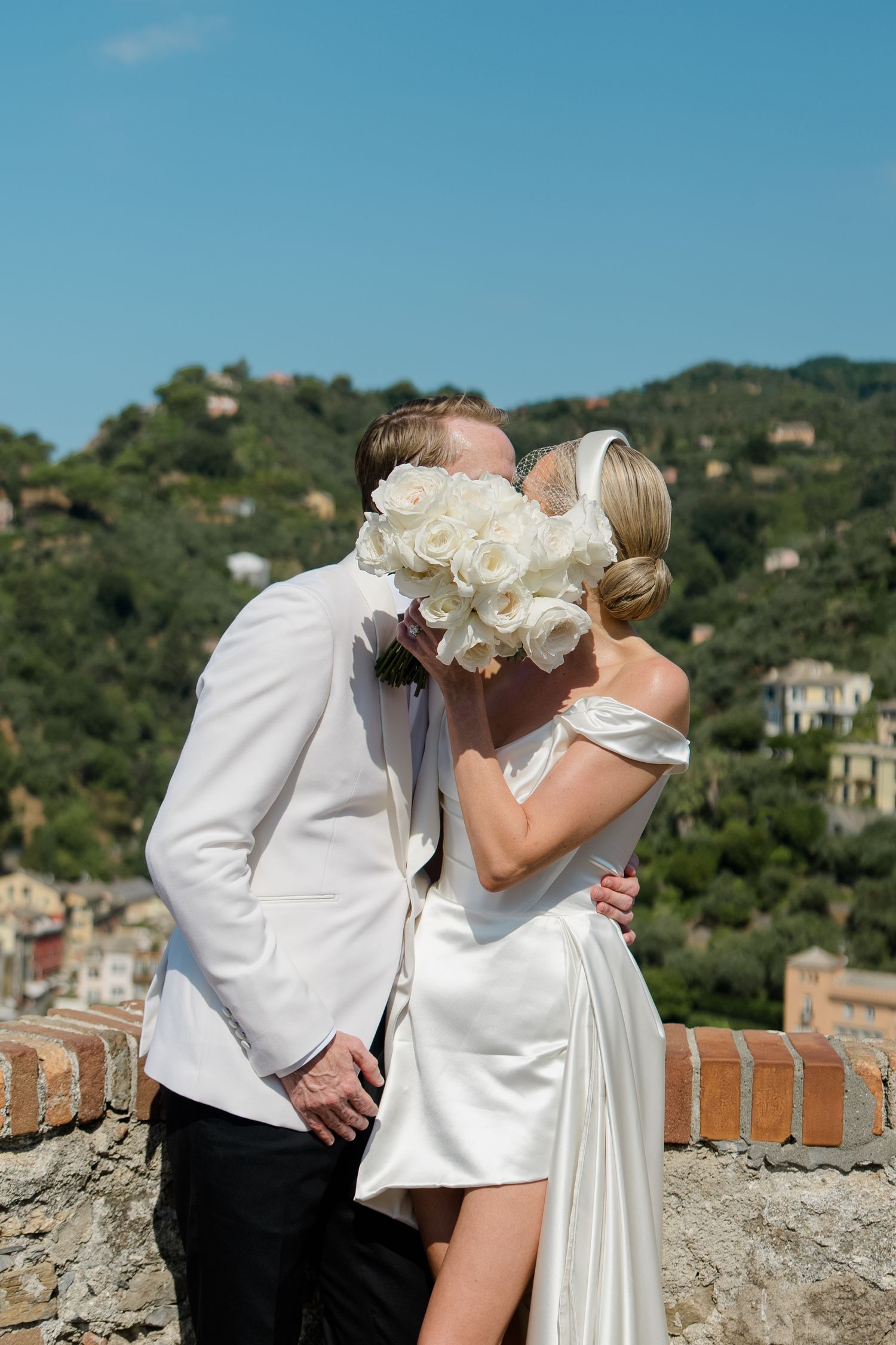 Bride and groom sharing a kiss hidden behind a white rose bouquet with a lush Italian hillside behind them.