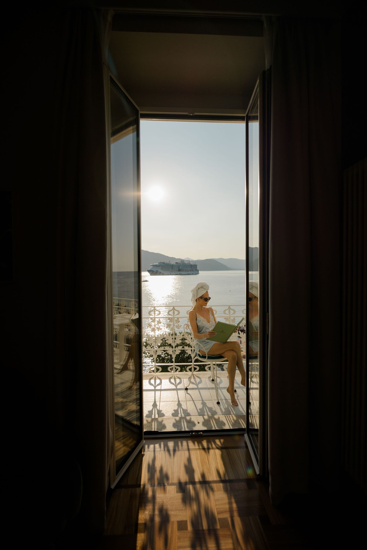 Woman in towel turban reads on sunlit balcony overlooking Mediterranean sea, viewed through open French doors.
