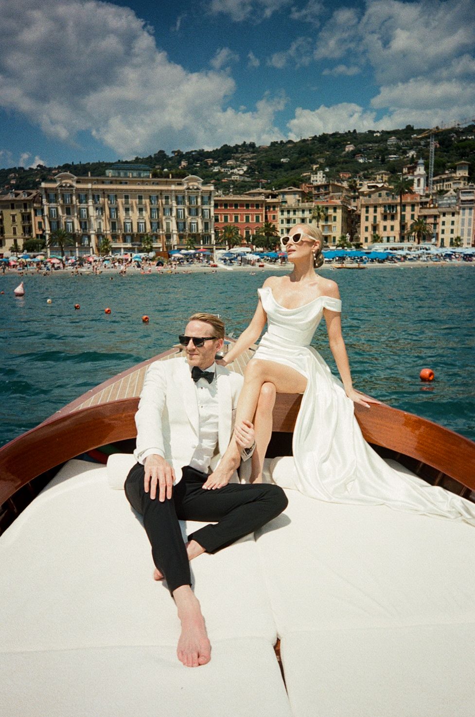 Bride in white gown perched on bow of wooden boat with groom in white tuxedo, Italian coastal town behind them.
