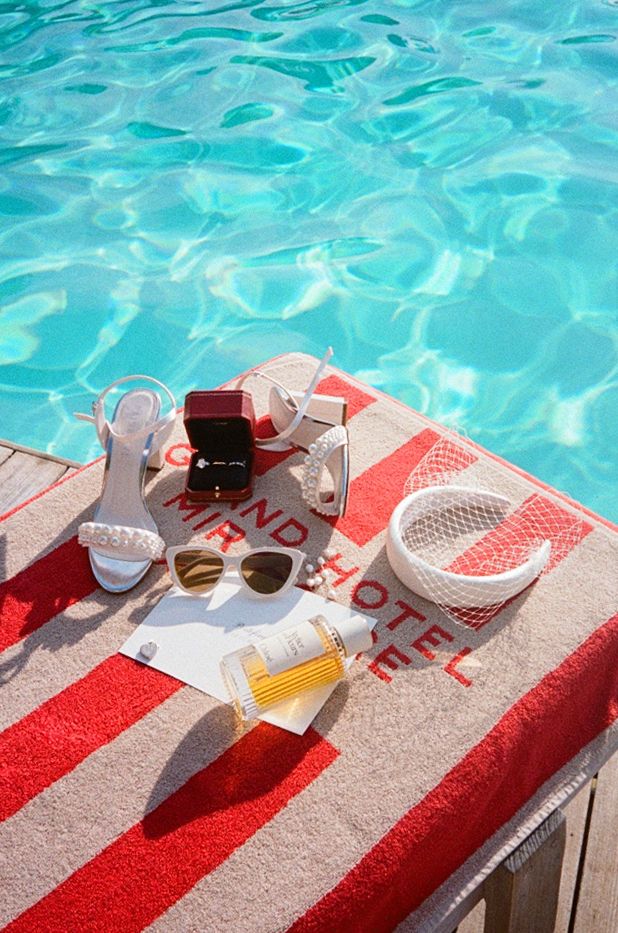 Grand Hotel pool towel with white heels, sunglasses, sunscreen, and accessories by a turquoise pool.