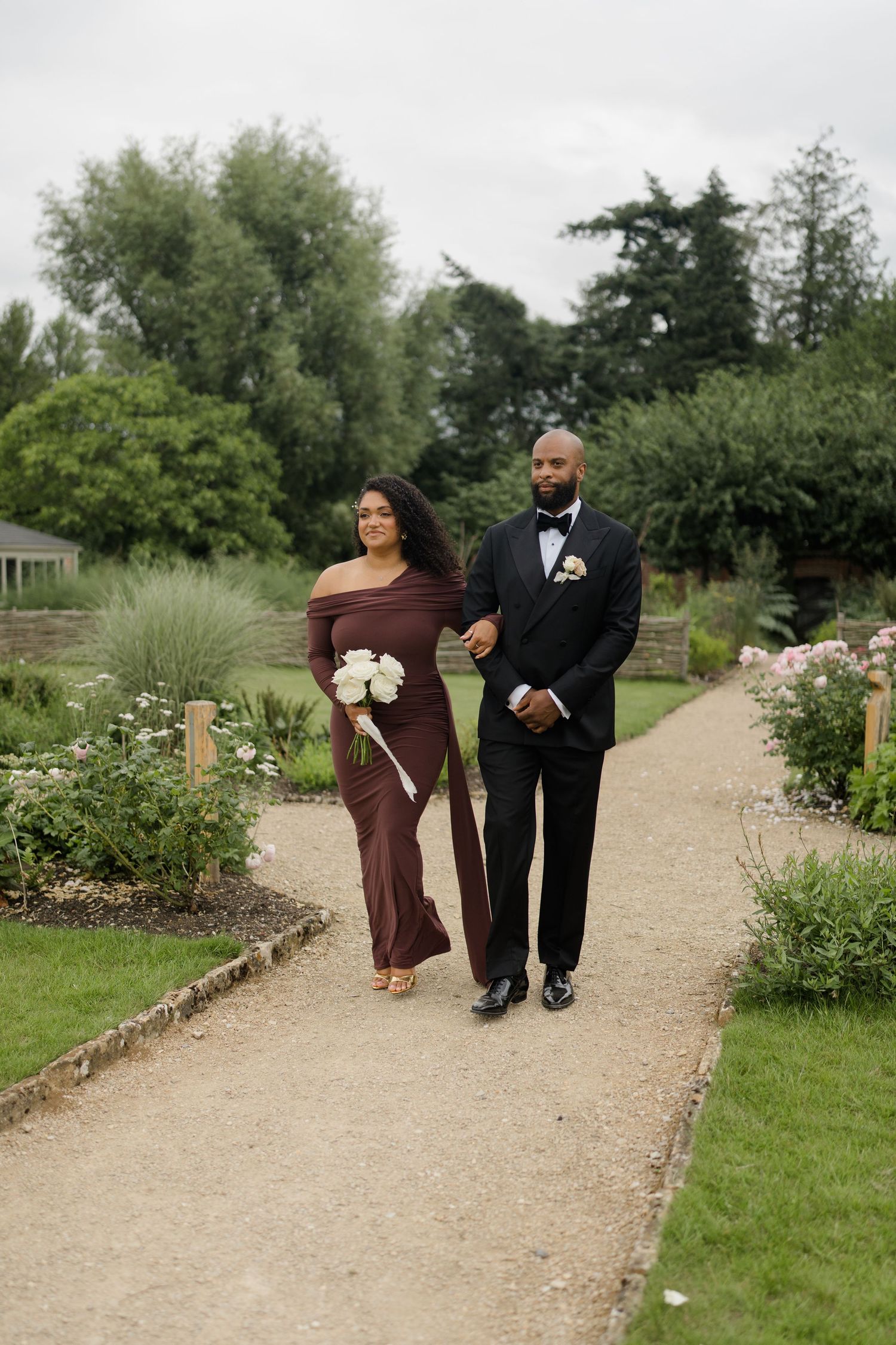 A bridesmaid in a burgundy off-shoulder gown walks with a groomsman in a black tuxedo along a garden path.