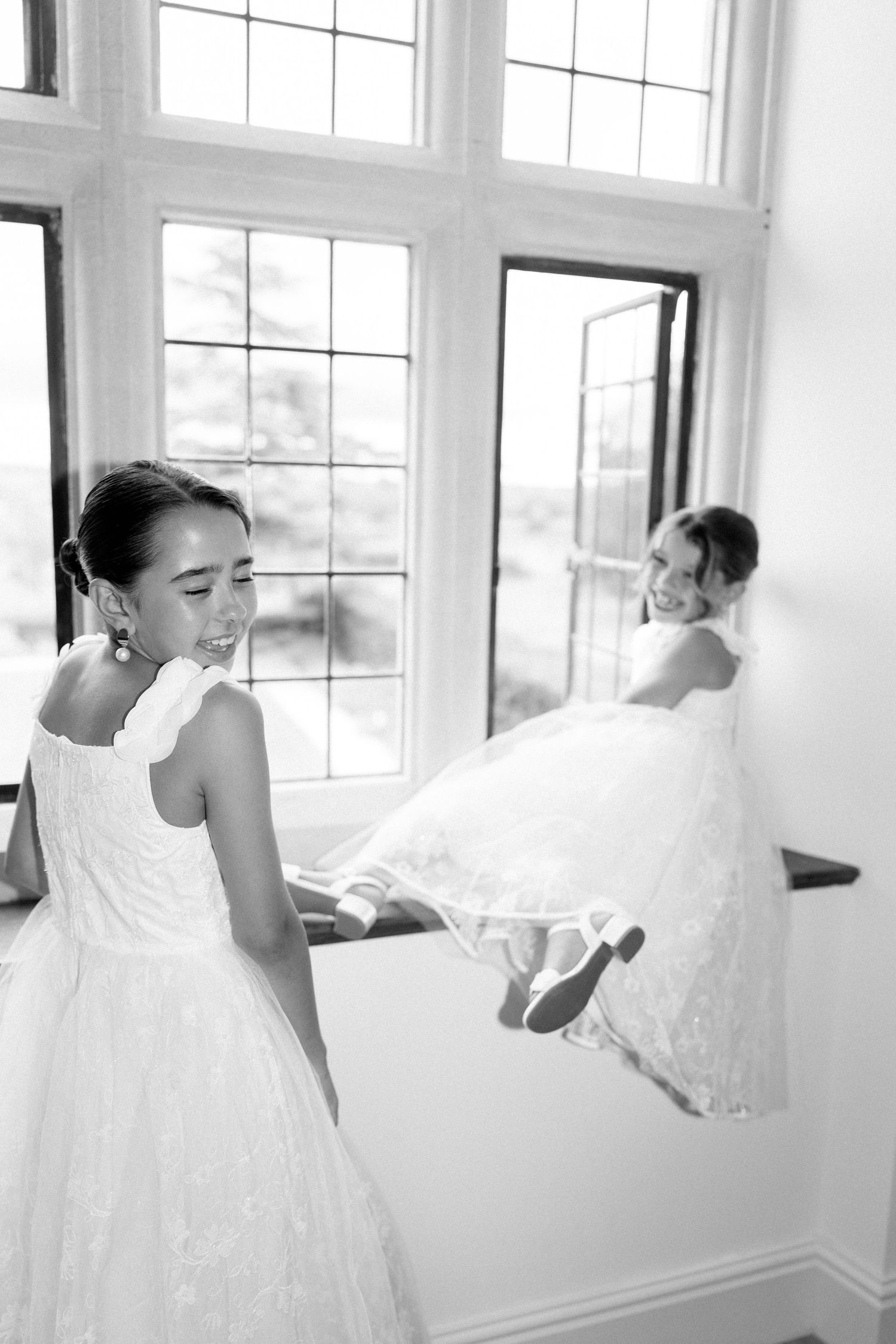 Two flower girls in white dresses playing near large windows in black and white.
