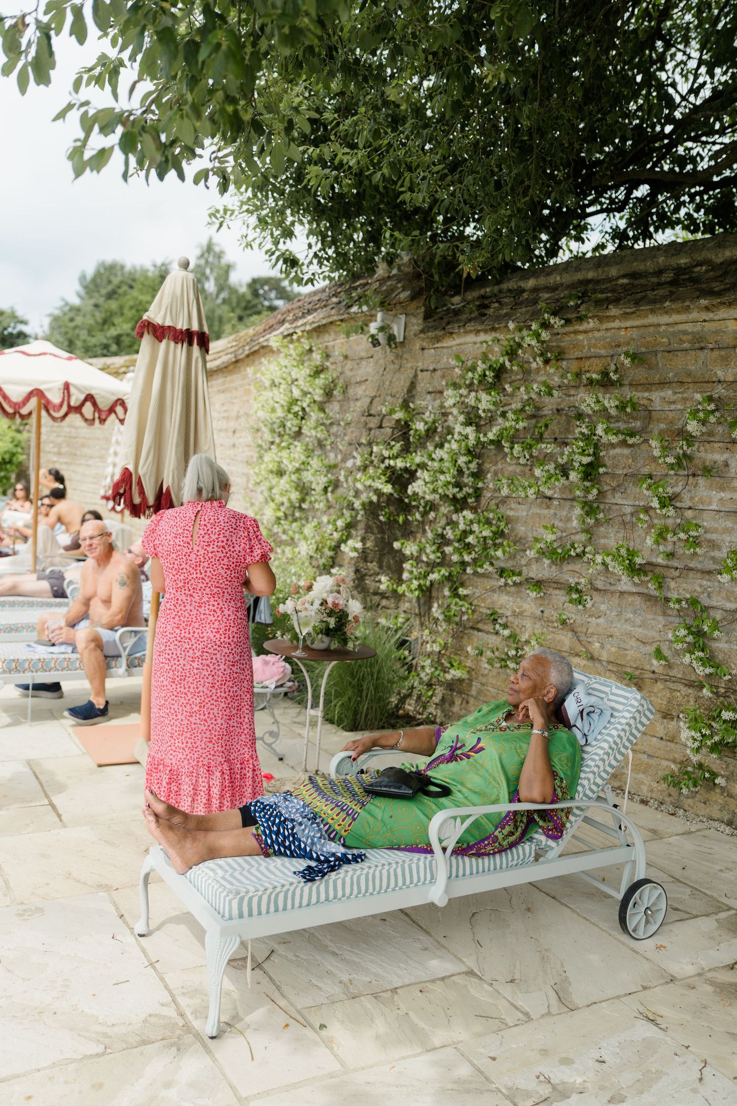 Elderly woman reclining on lounge chair beside stone wall at outdoor wedding garden party.