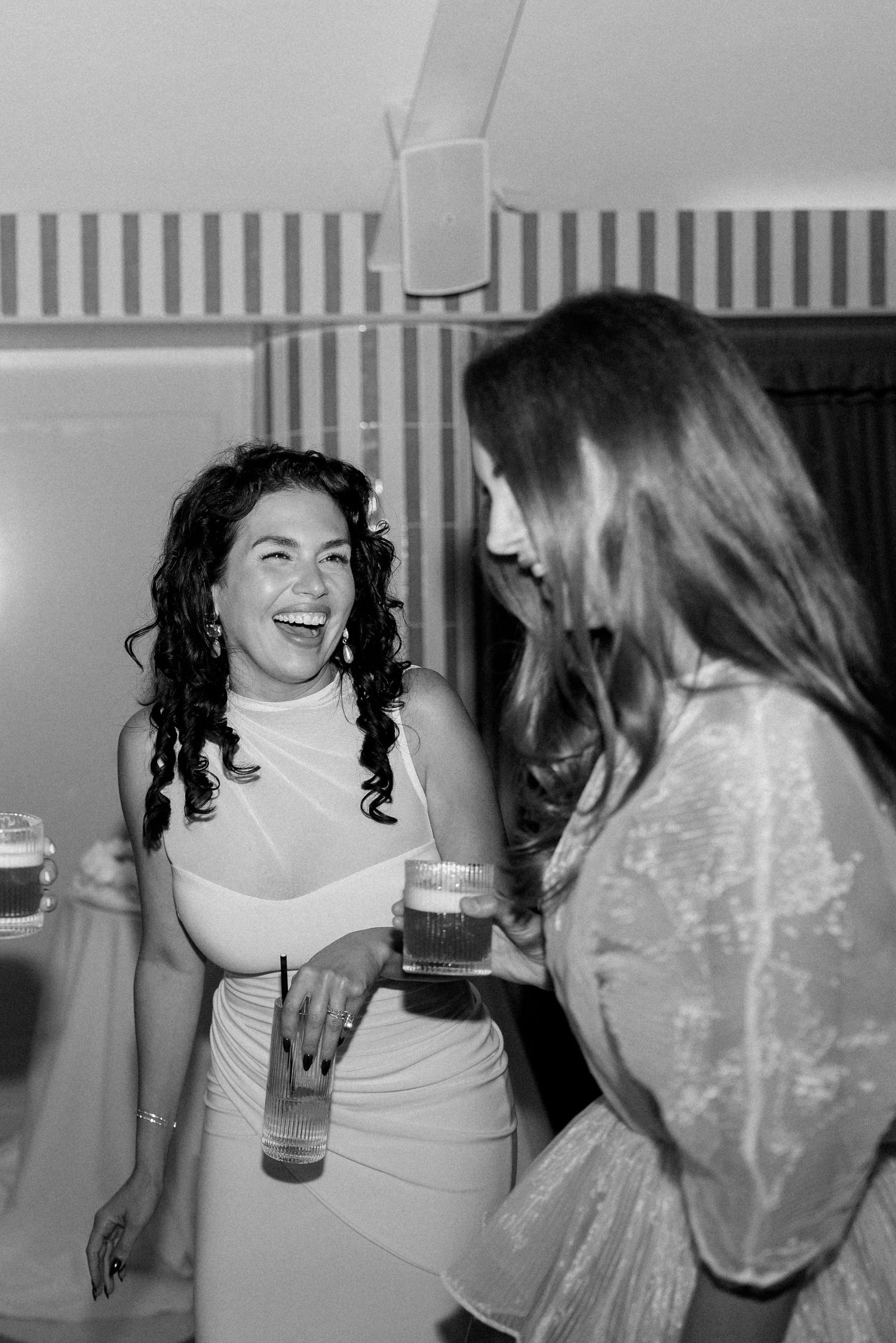 Two women laughing and holding drinks at lively black and white wedding reception party.