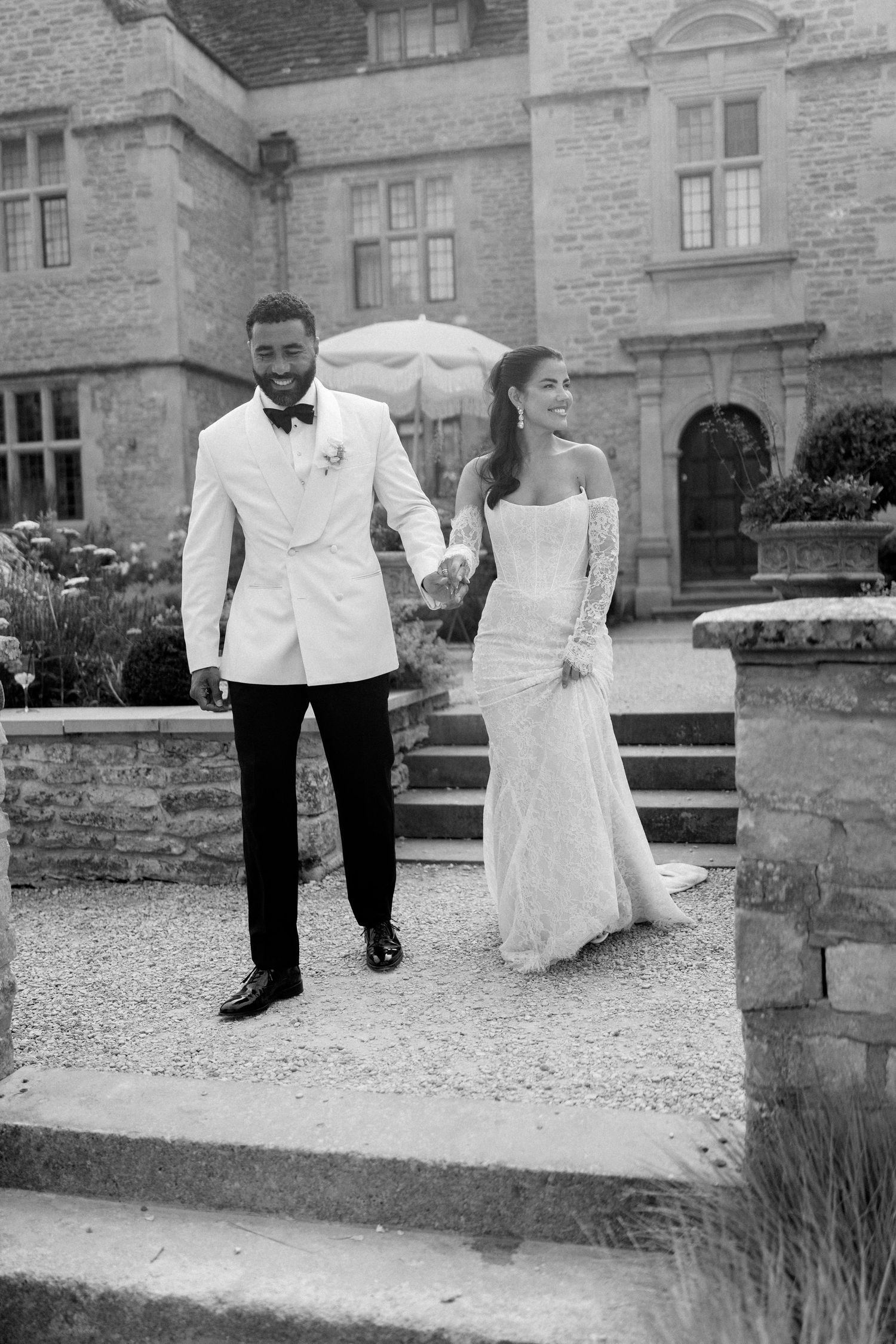 Black and white photo of elegant bride and groom holding hands walking outside historic stone manor.