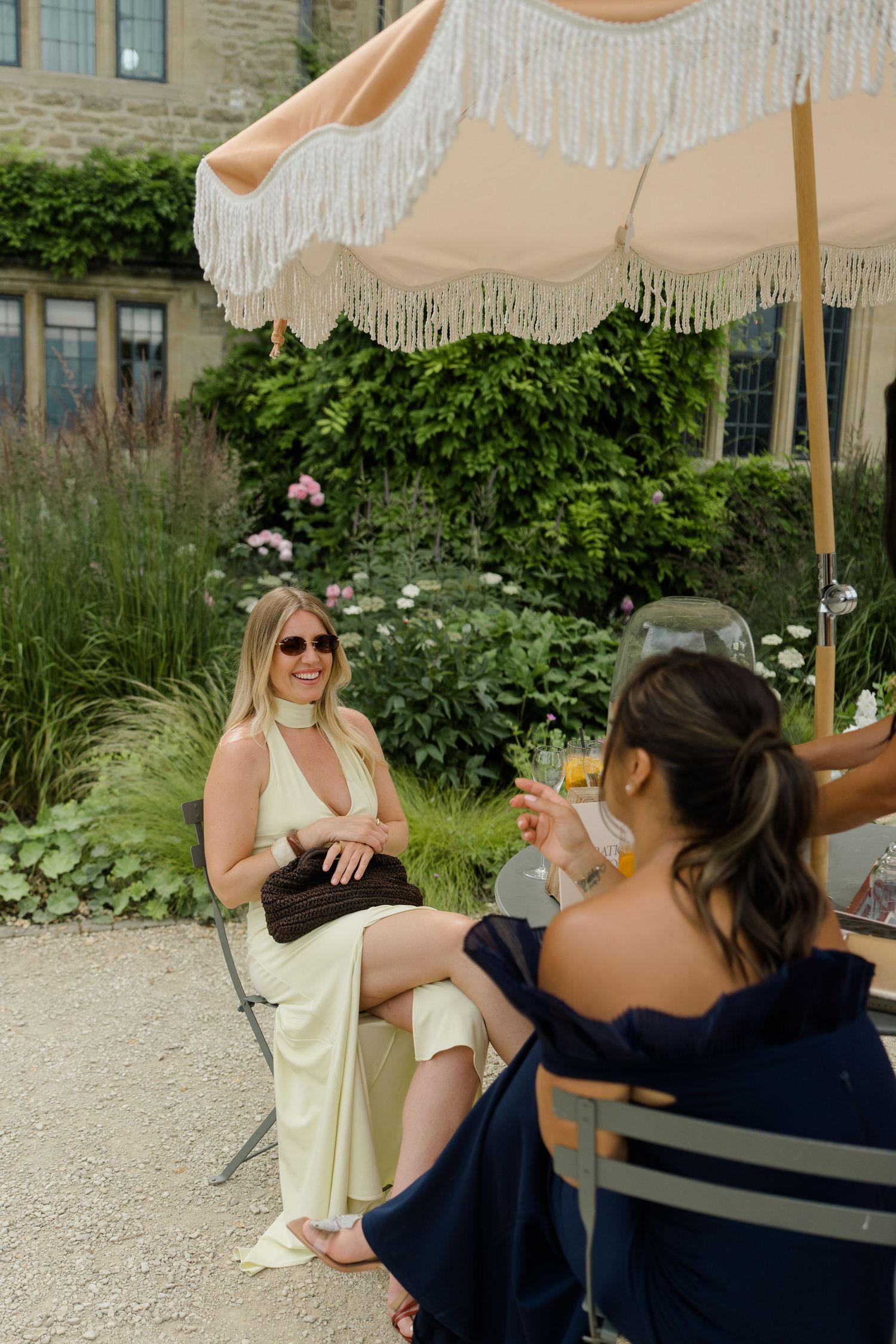 Two elegantly dressed women relax under a fringed parasol at an outdoor wedding reception in a garden setting.