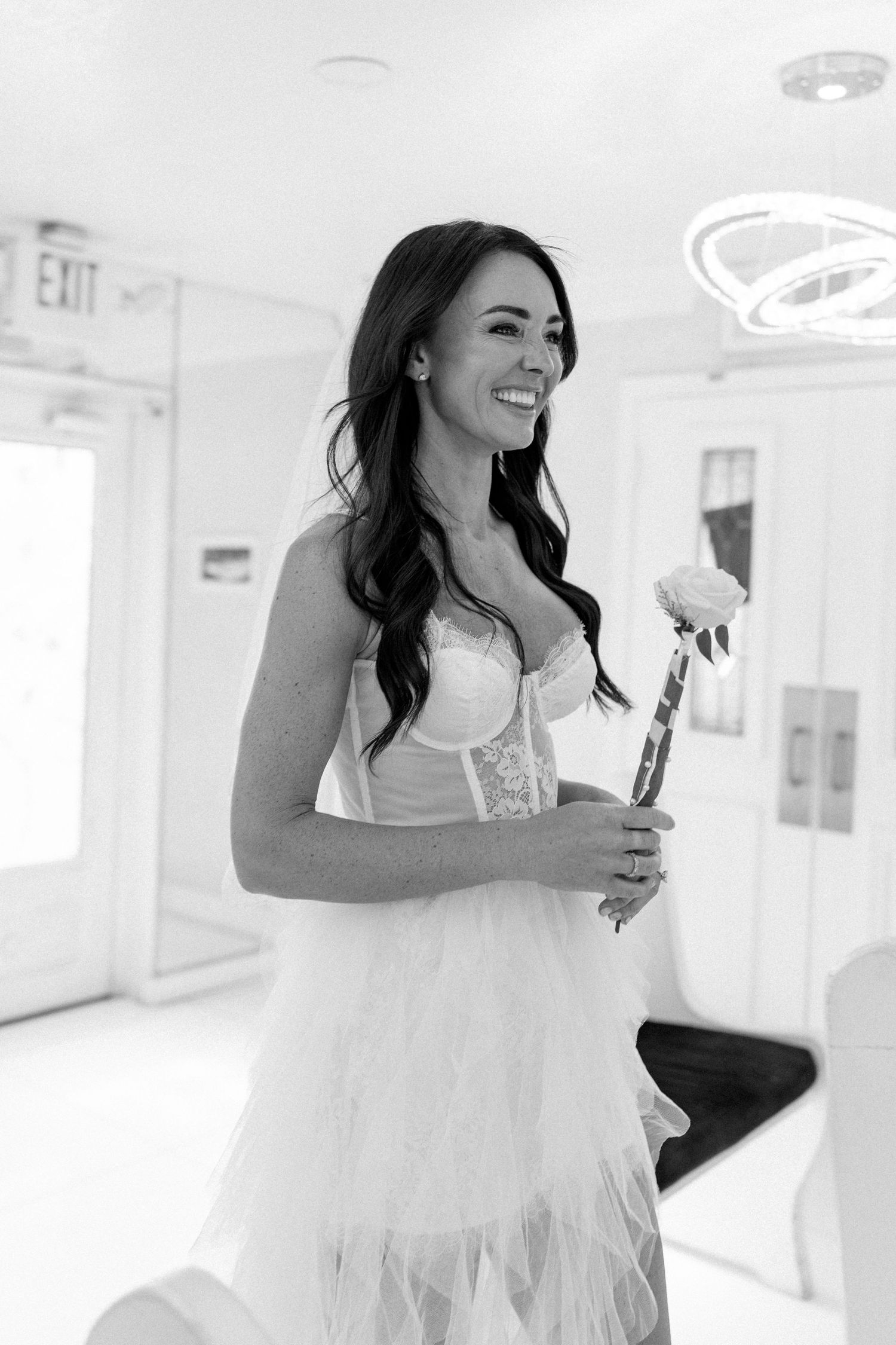 A bride in a white corset dress holds a rose while smiling in a bright indoor venue.