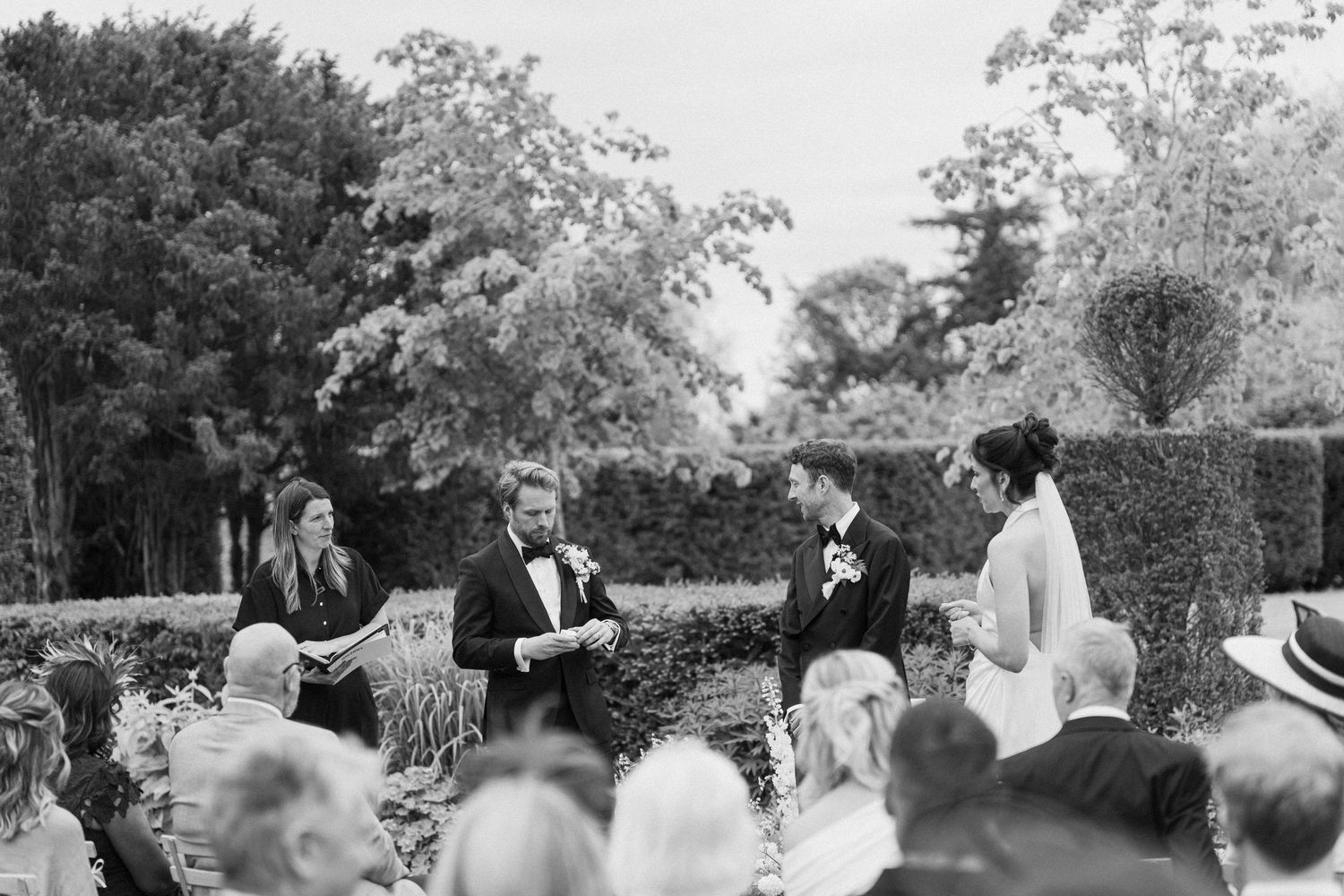 Black and white photo of outdoor garden wedding ceremony with couple exchanging vows before seated guests.