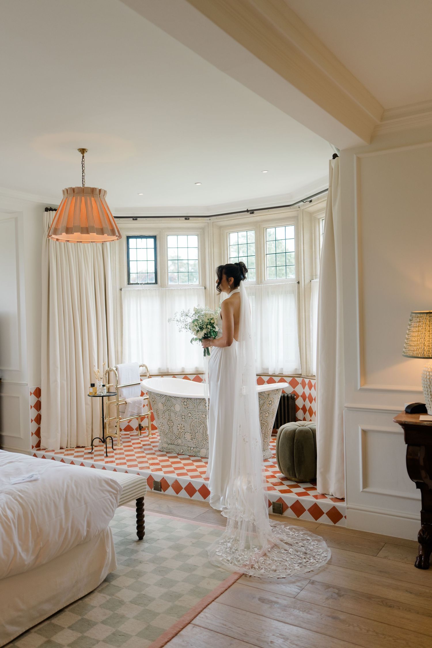 Bride in white gown holding bouquet stands by bay window with freestanding bathtub in elegant bridal suite.
