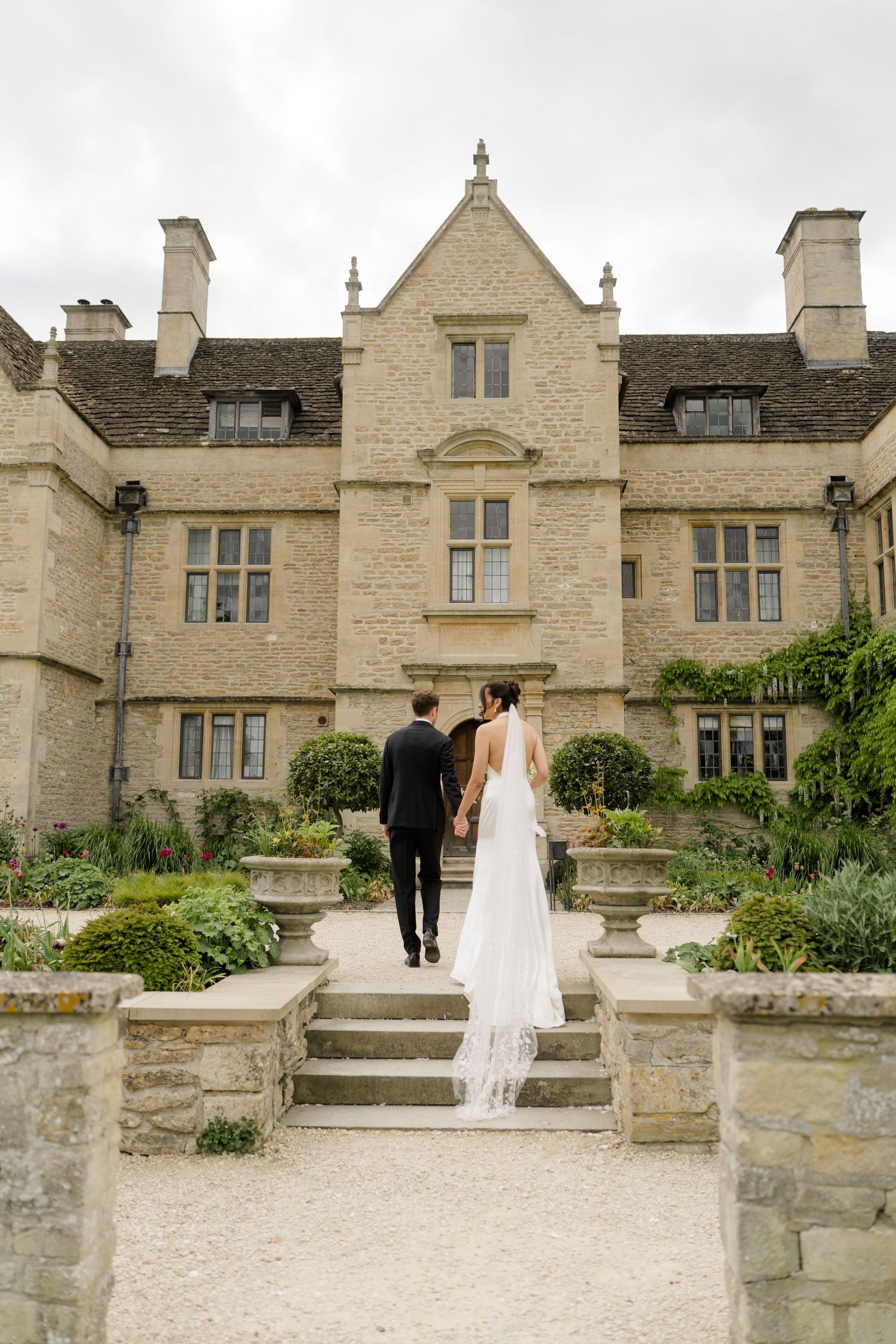 Bride in cathedral veil and groom walk hand-in-hand toward grand Cotswold stone manor wedding venue.