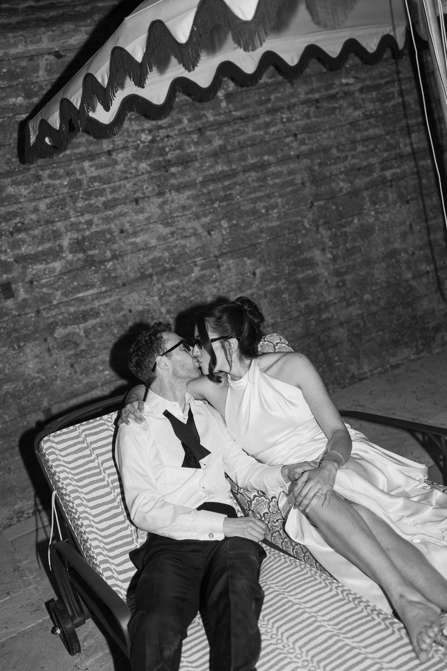 Black and white romantic photo of bride and groom kissing on lounge chair under striped awning.