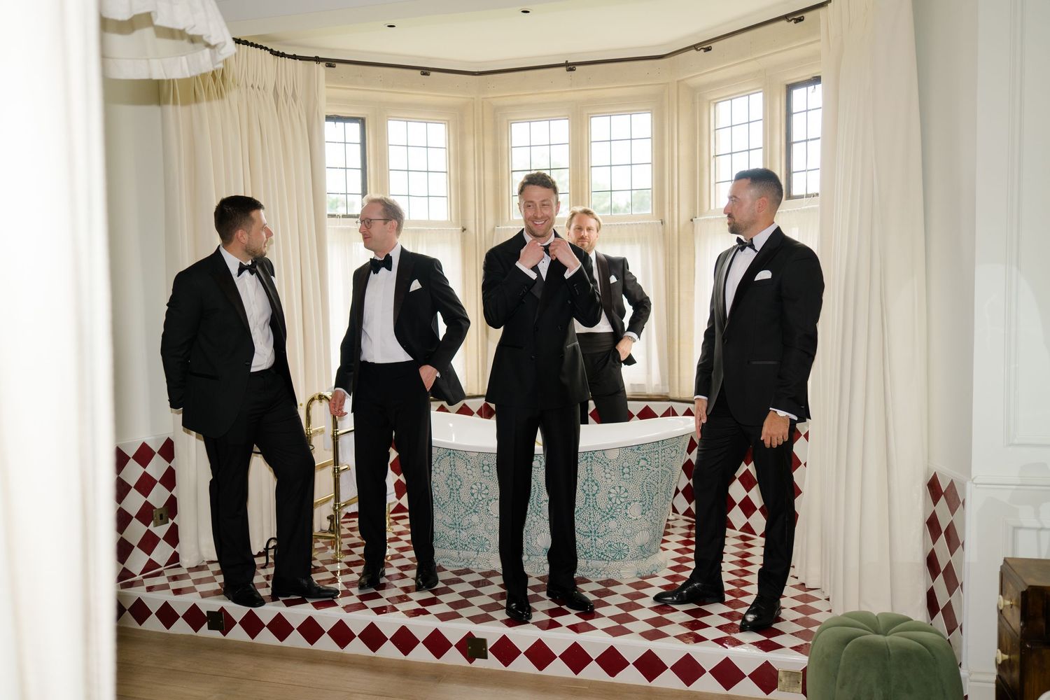Five groomsmen in black tuxedos gathered in elegant bathroom with red checkered tile floor.