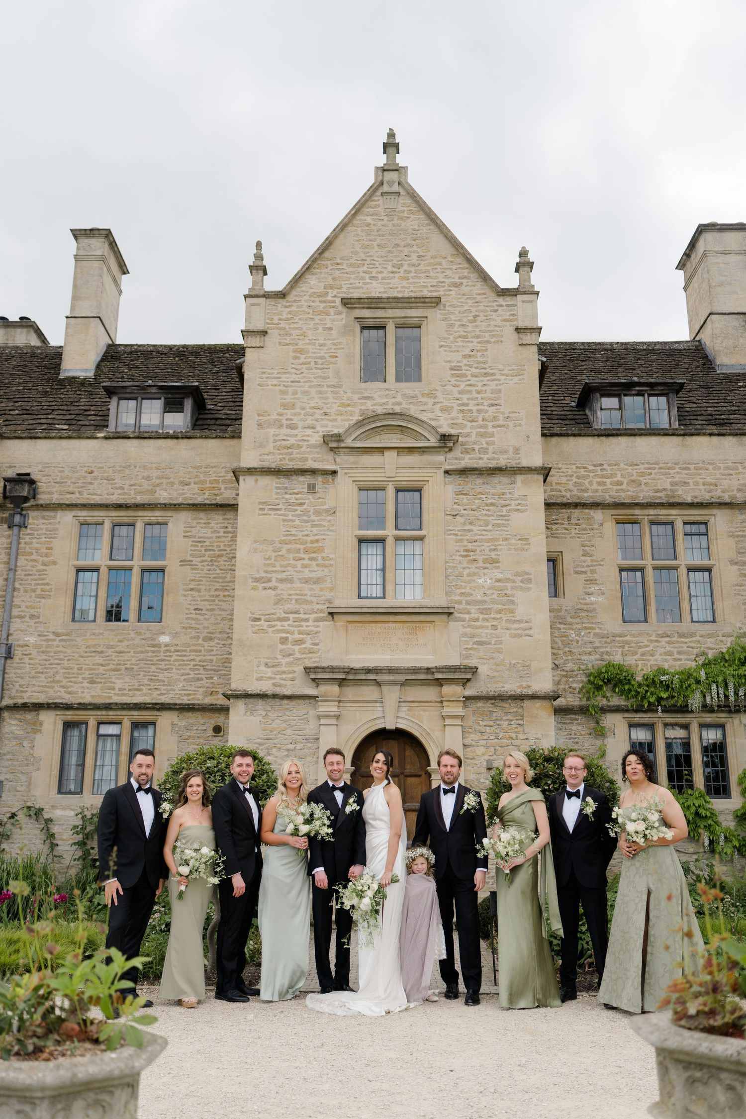 Wedding party poses in front of grand Cotswolds stone manor, bridesmaids in sage green gowns with groomsmen in black tuxedos.