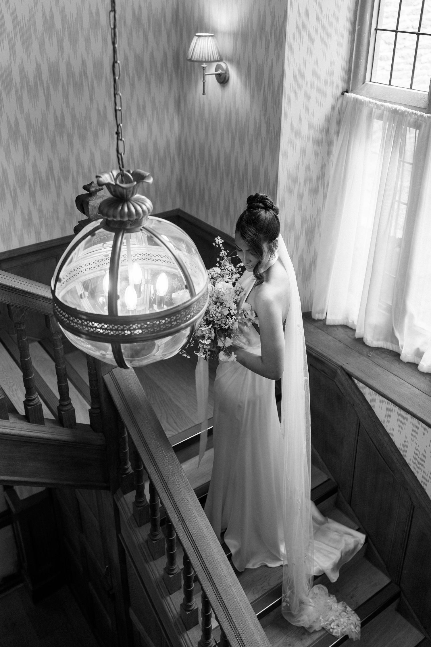 Black and white aerial view of bride in gown descending ornate wooden staircase with bouquet.