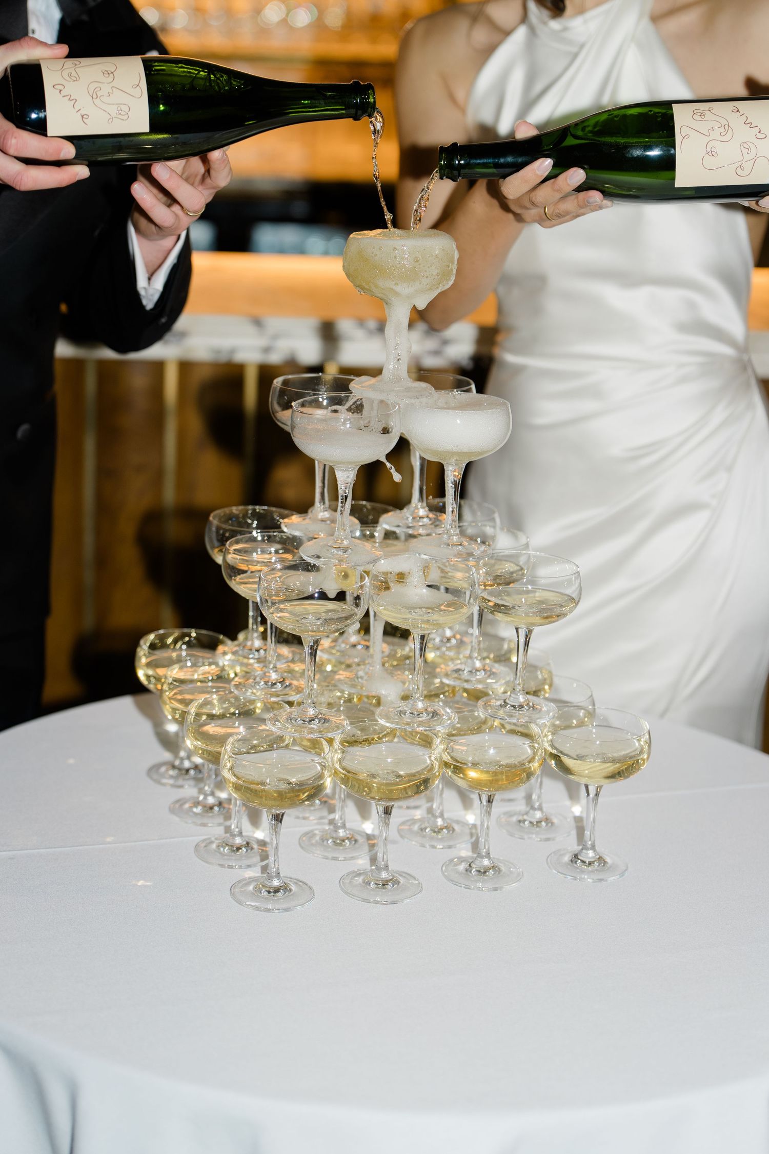 Champagne tower being filled at wedding reception with bride in white gown pouring bubbly.