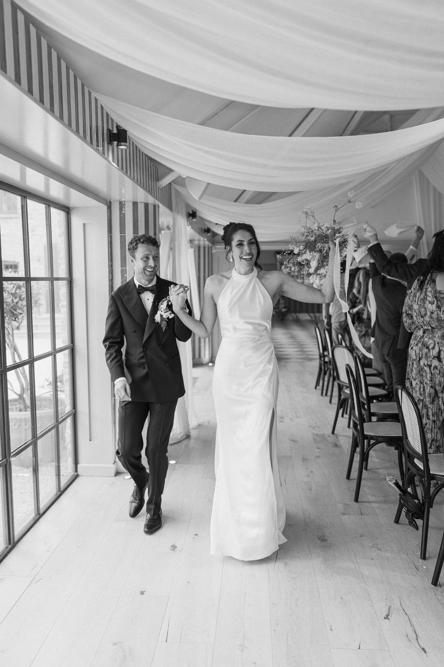 Joyful bride and groom make grand entrance into elegant draped wedding reception venue in black and white photo.